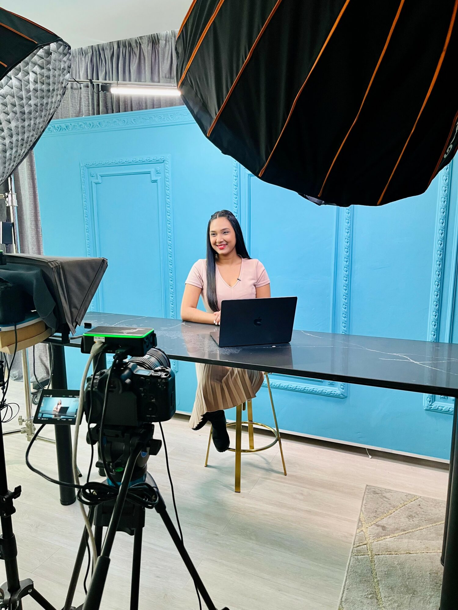 Woman sitting at a desk with a laptop, in a studio with blue walls and professional lighting equipment.