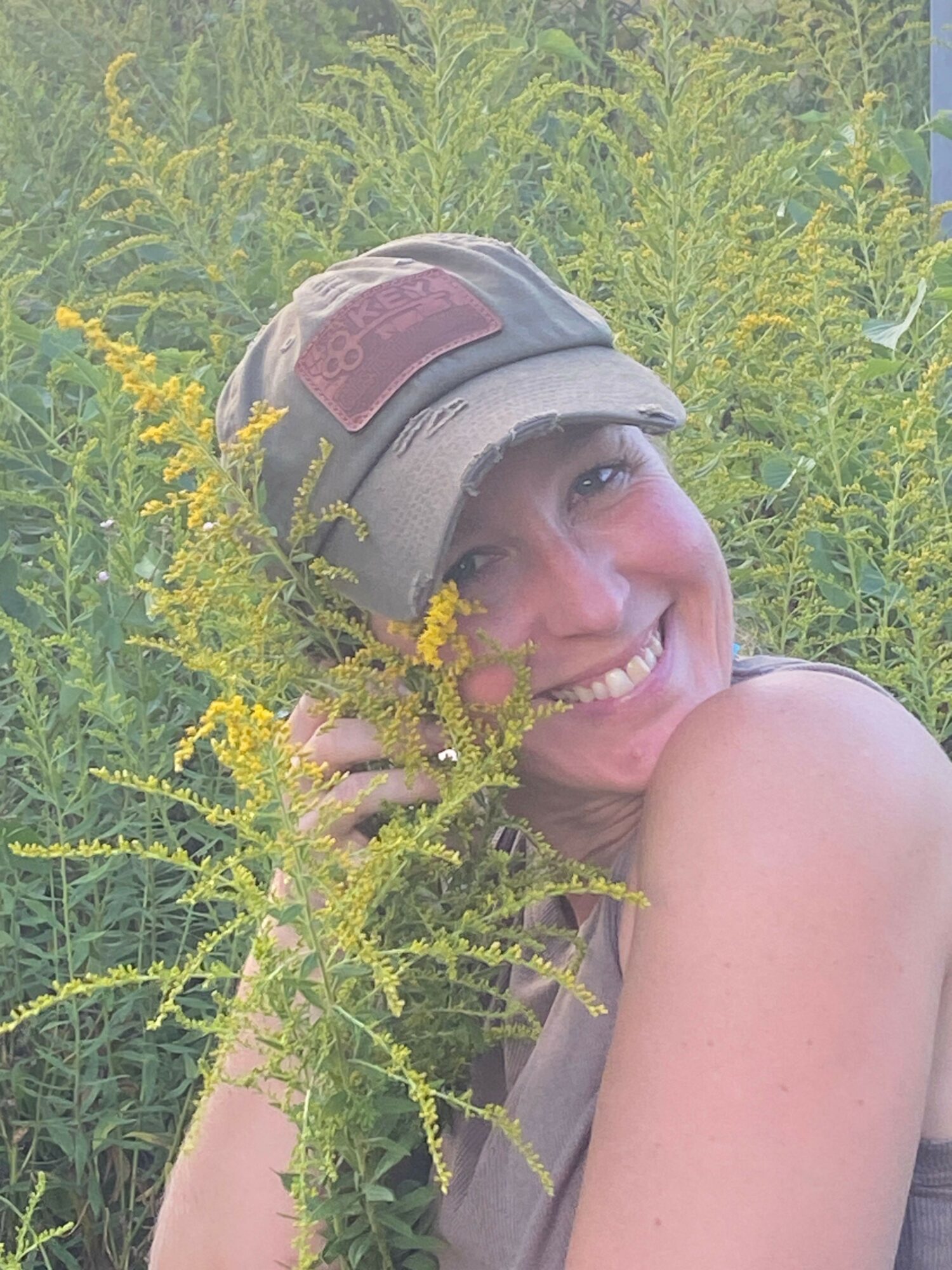 Person lying in grass with a helmet, smiling, surrounded by yellow flowers and green plants.