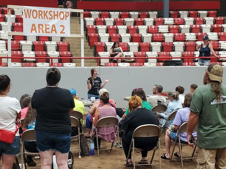 Person speaking to seated audience in an outdoor workshop area with red and white stadium seats in background.