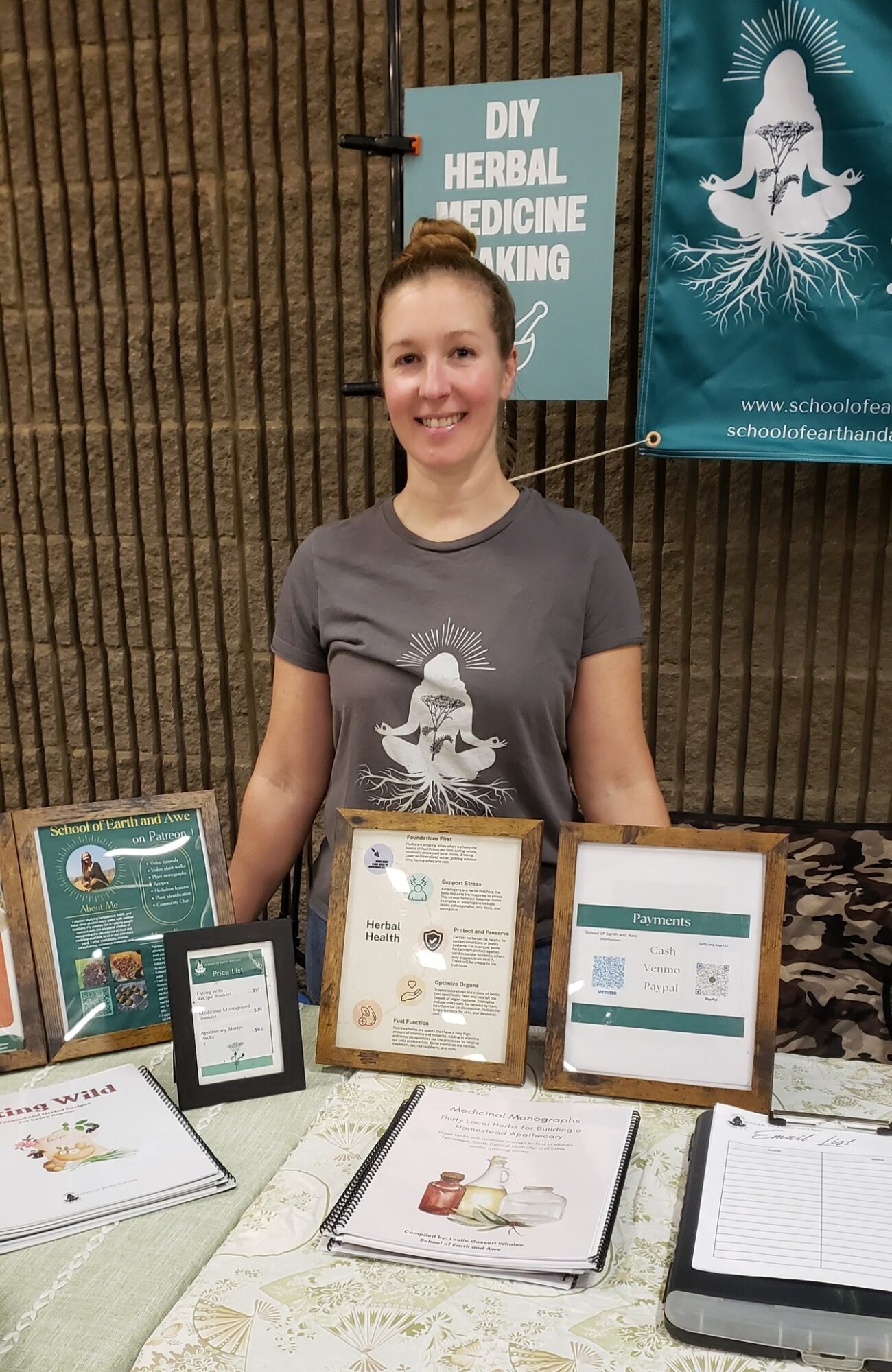 Smiling woman standing behind a table with herbal medicine information displays and framed certificates, outdoors with a wooden fence background.