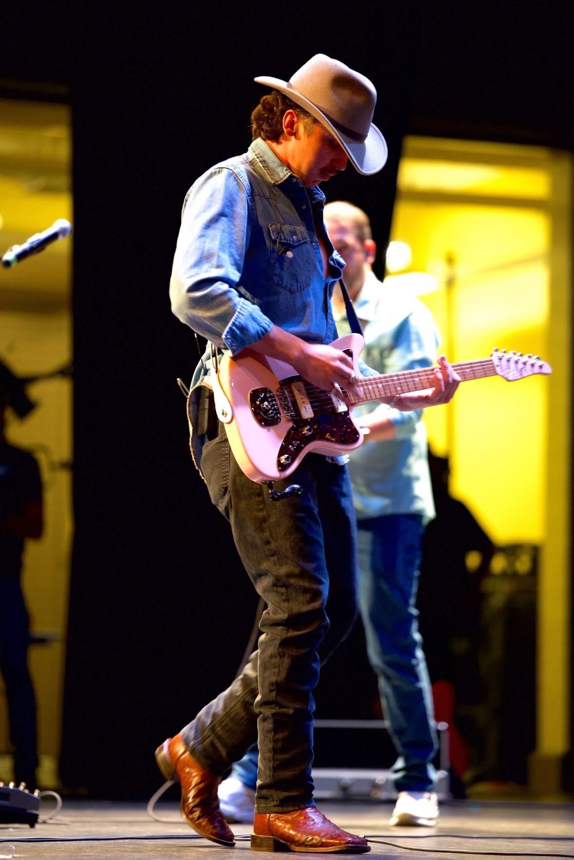Young musician playing pink electric guitar on stage with yellow background, wearing a hat, denim shirt, and brown boots.