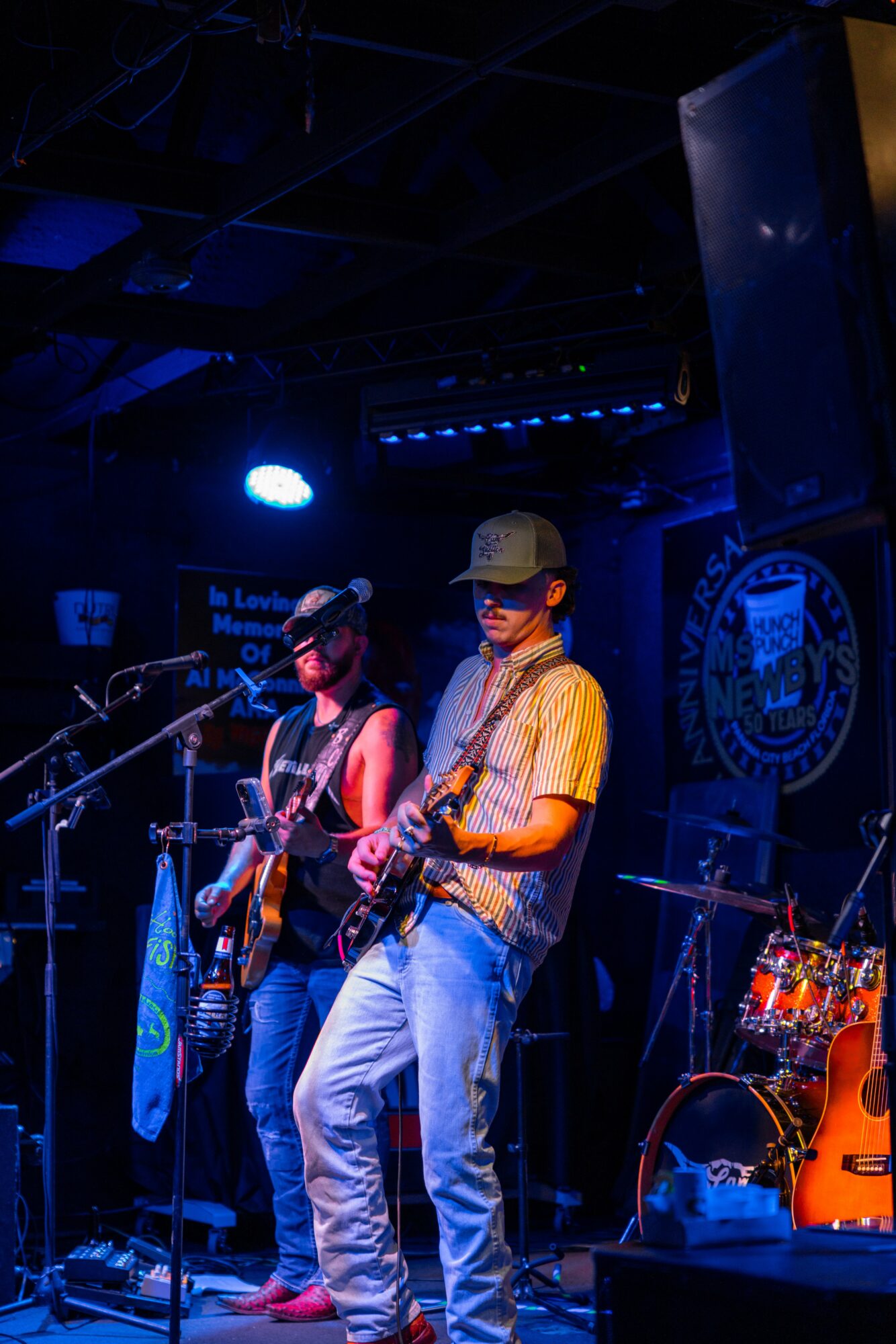 Two musicians perform on stage with guitars, wearing casual clothes and hats, under blue stage lighting.