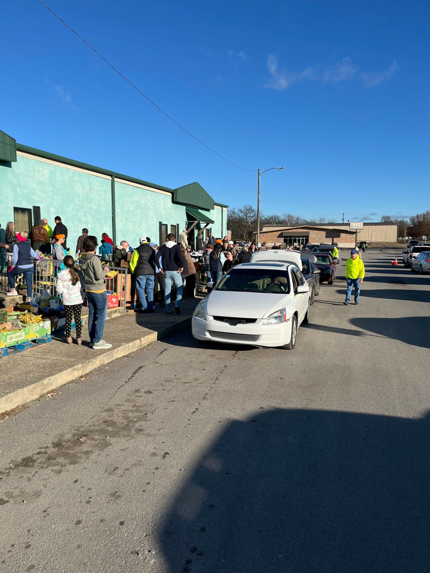 People gather outside a building with a light green roof, with cars parked nearby and a clear blue sky.