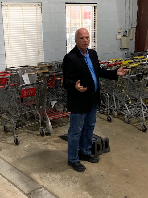 Man in a black jacket and blue shirt standing in a store aisle with shopping carts, gesturing with hands.