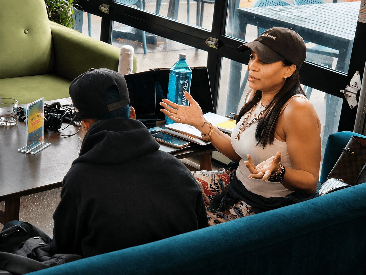 Two people sitting at a table in a cafe, one woman speaking and gesturing, the other person listening, with a large window behind them.