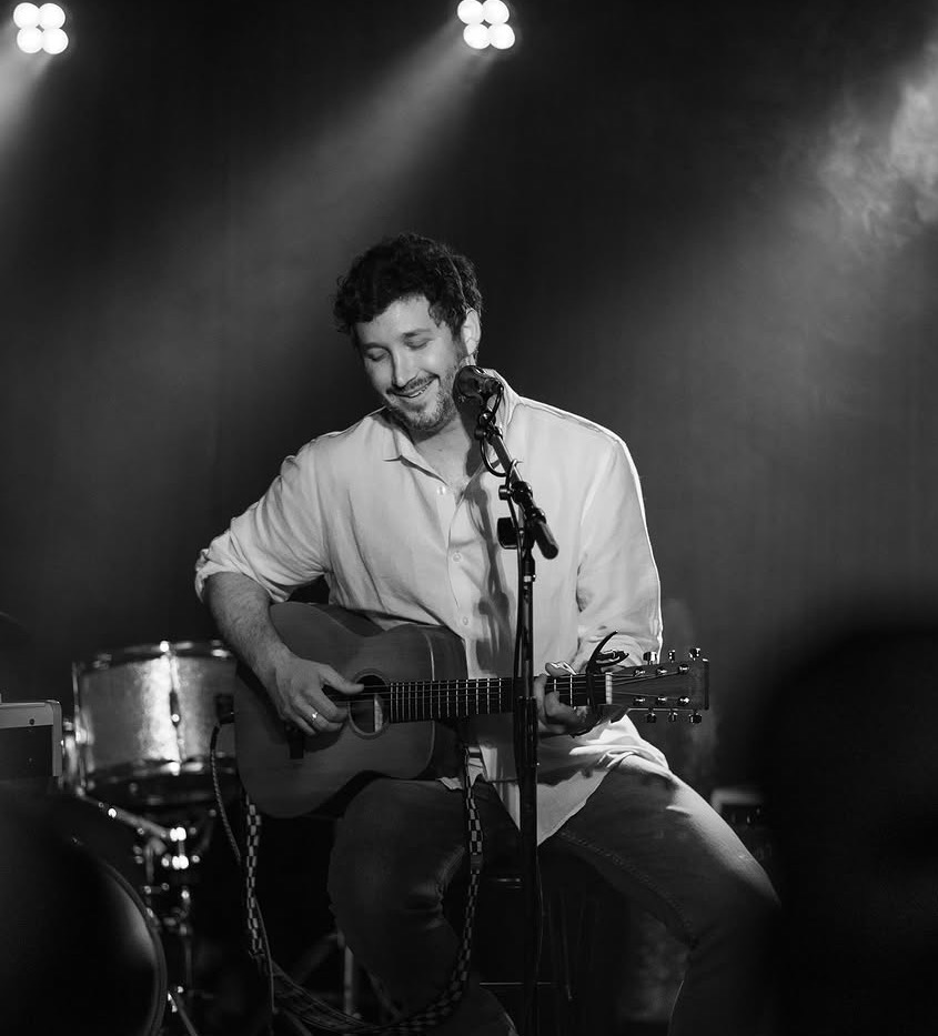 Man playing guitar and singing into microphone on stage with drums in background, black and white image.