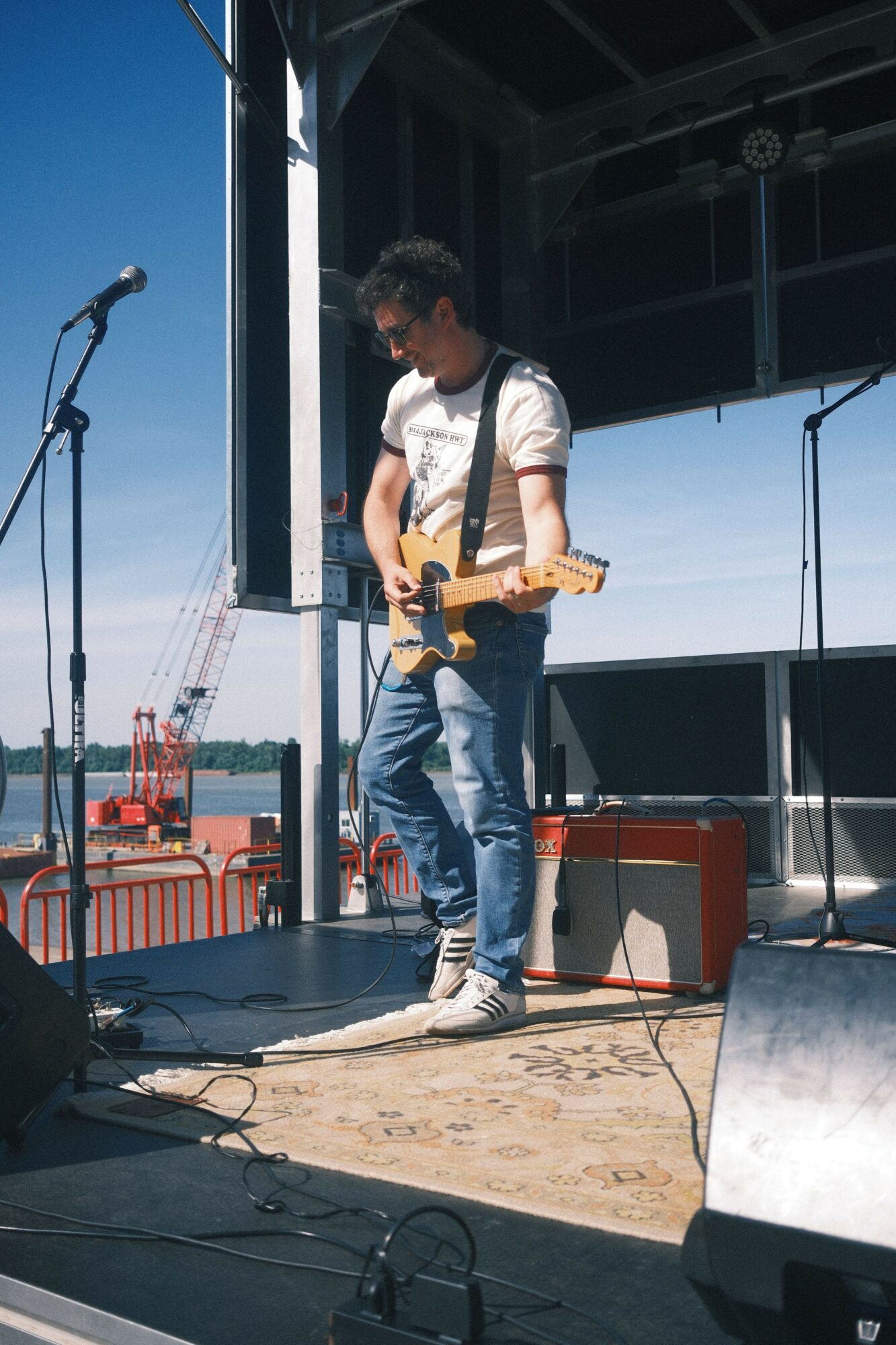 Young man standing on a rooftop with city skyline in background, holding a wooden object, wearing jeans and a white shirt.