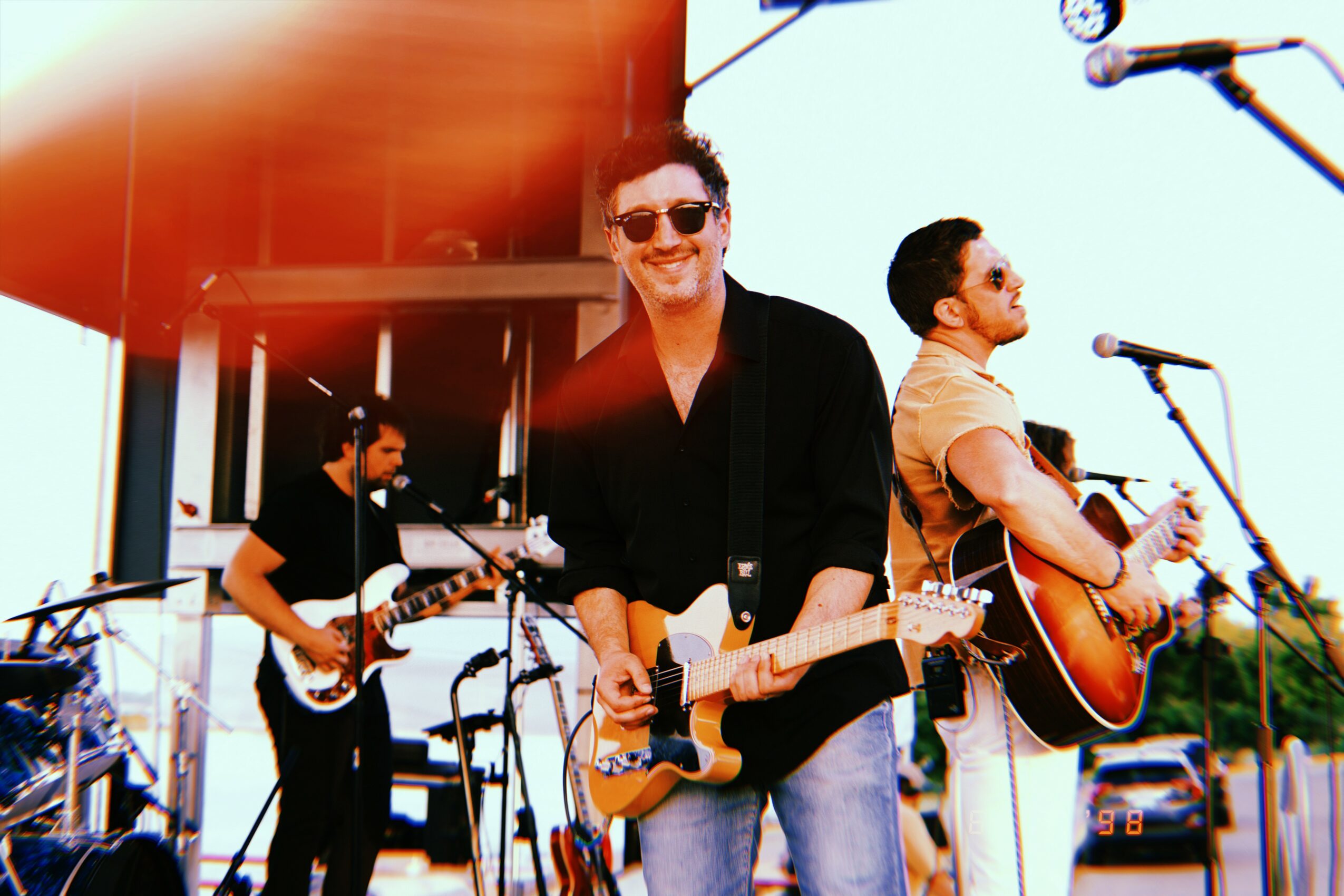 Three musicians perform outdoors with guitars, microphones, and equipment, smiling and enjoying the music.