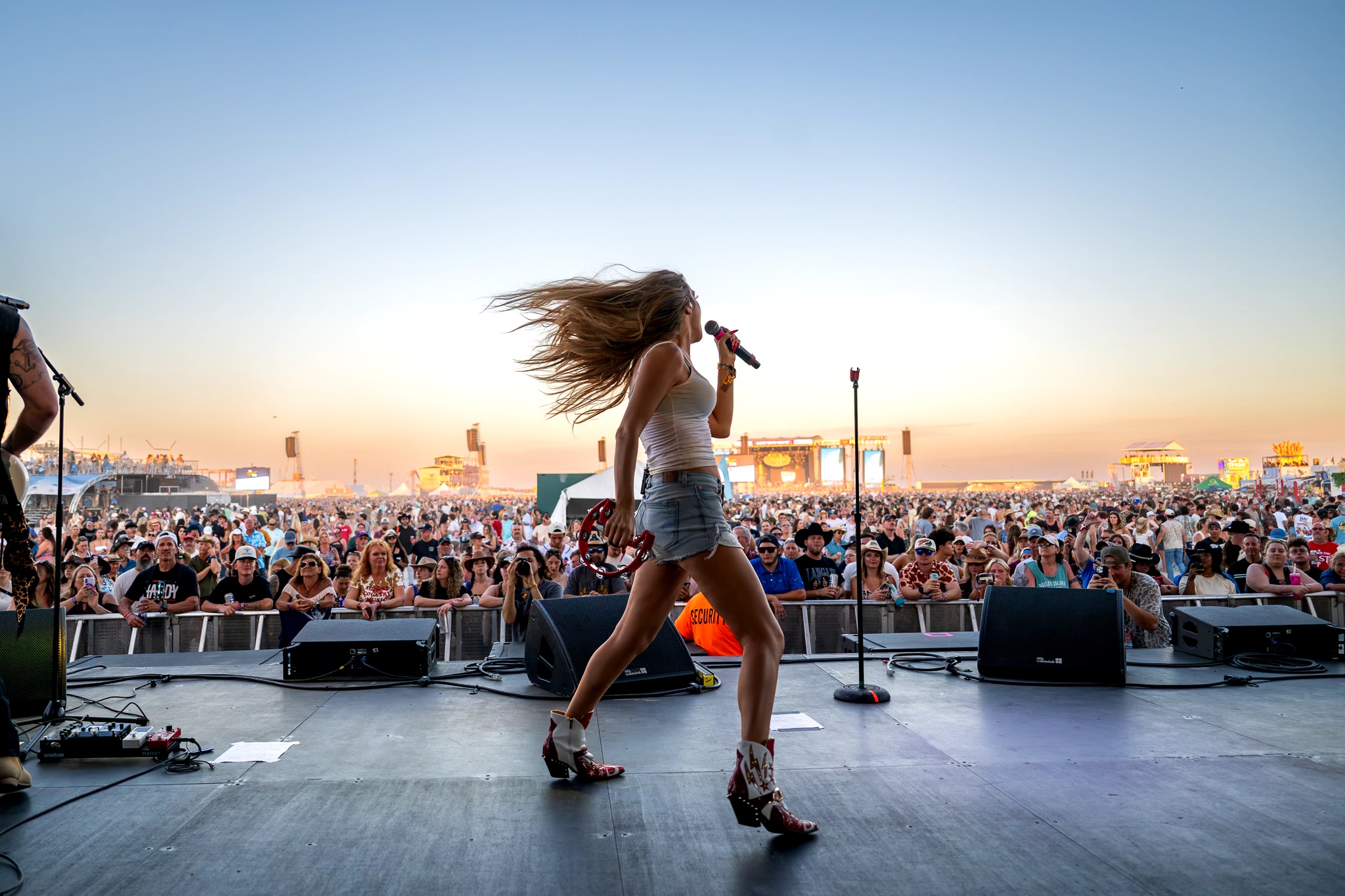 Performer with long hair dancing on stage during outdoor concert at sunset, crowd in background, holding microphone, stage equipment visible.