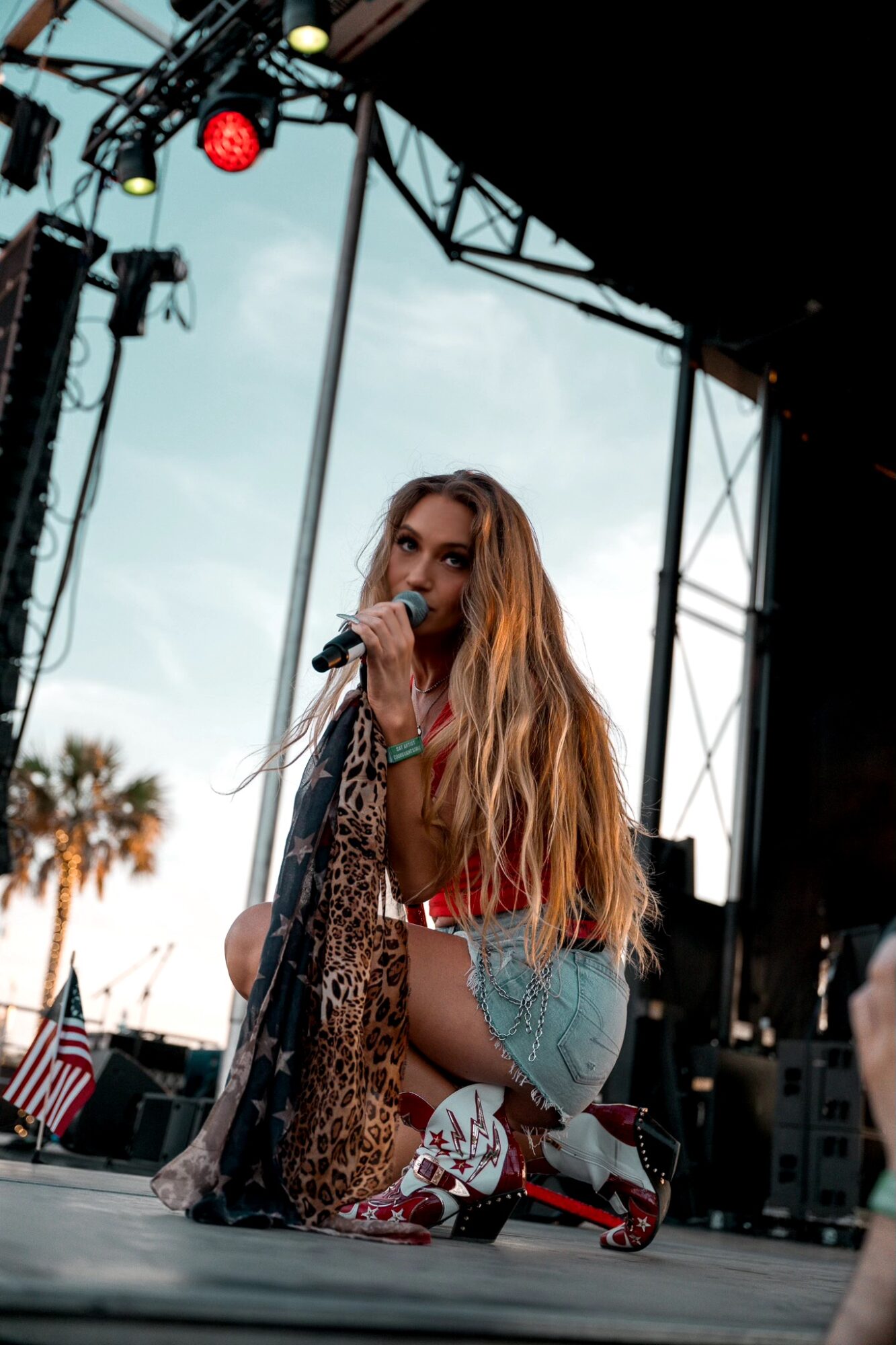 A woman with long hair holding a microphone on an outdoor stage, with stage equipment and sky visible behind her.