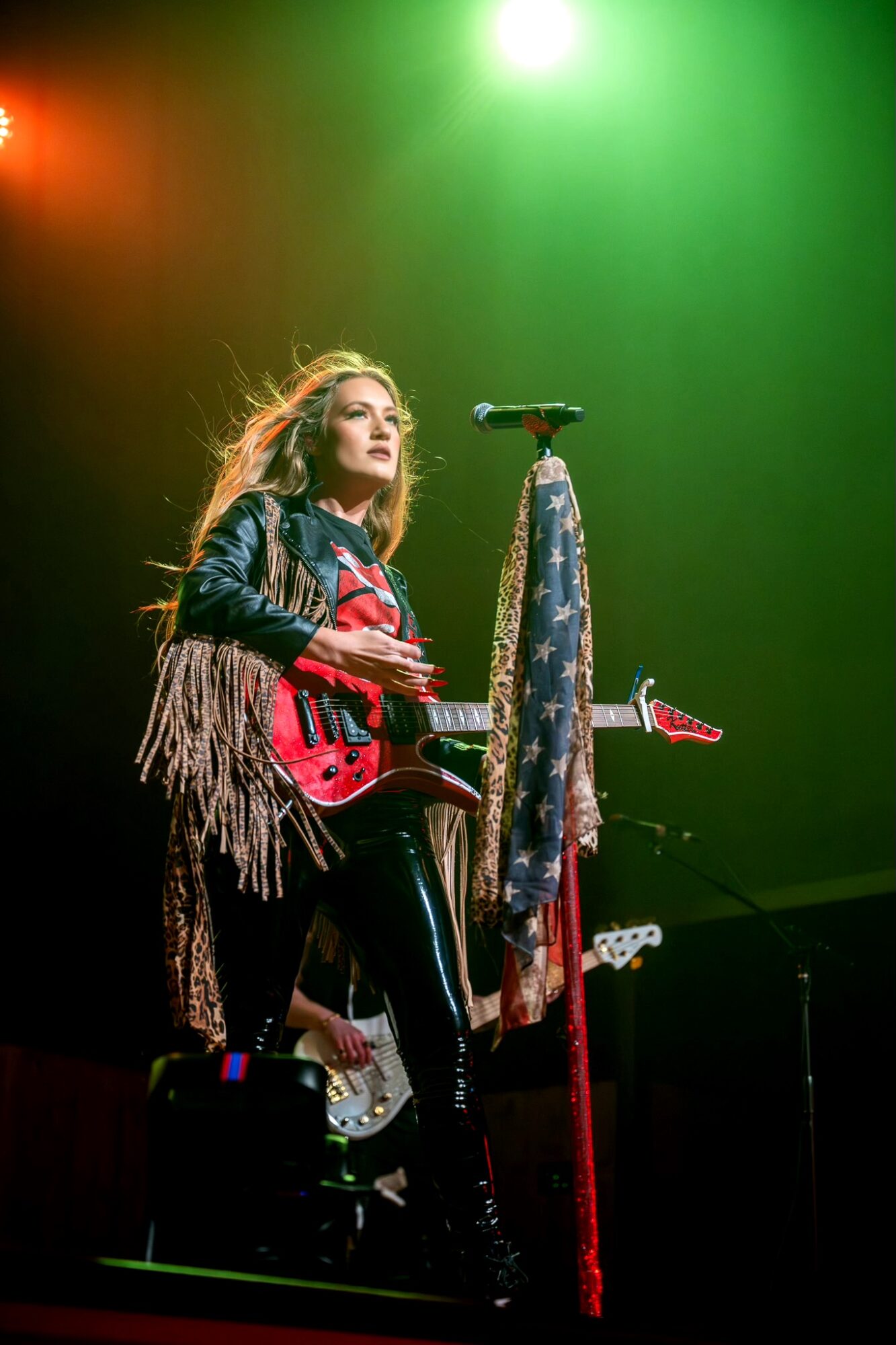 Female musician on stage with a guitar, microphone, and colorful stage lighting, performing live.