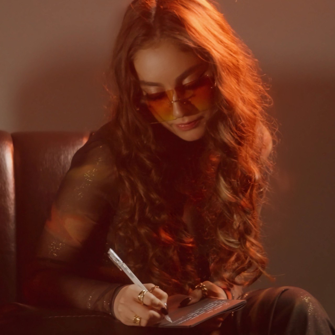 Woman with long curly hair wearing sunglasses and a black outfit, sitting and writing on a notepad.