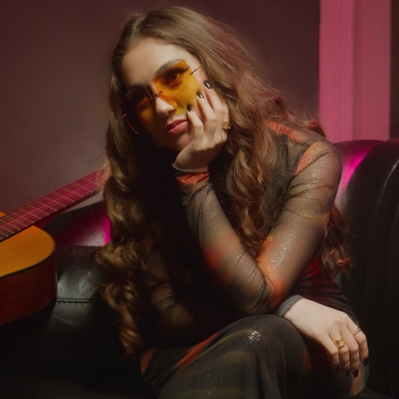 Young woman with long wavy hair wearing yellow-tinted sunglasses, sitting on a black leather couch with pink curtains behind her.