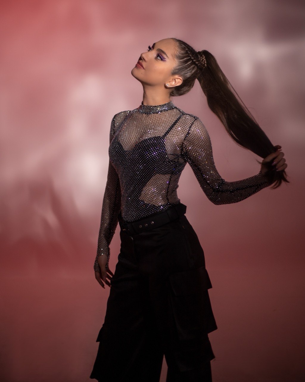 A woman with a high ponytail wearing a sheer, sparkly top and black pants, looking upward against a pinkish background.