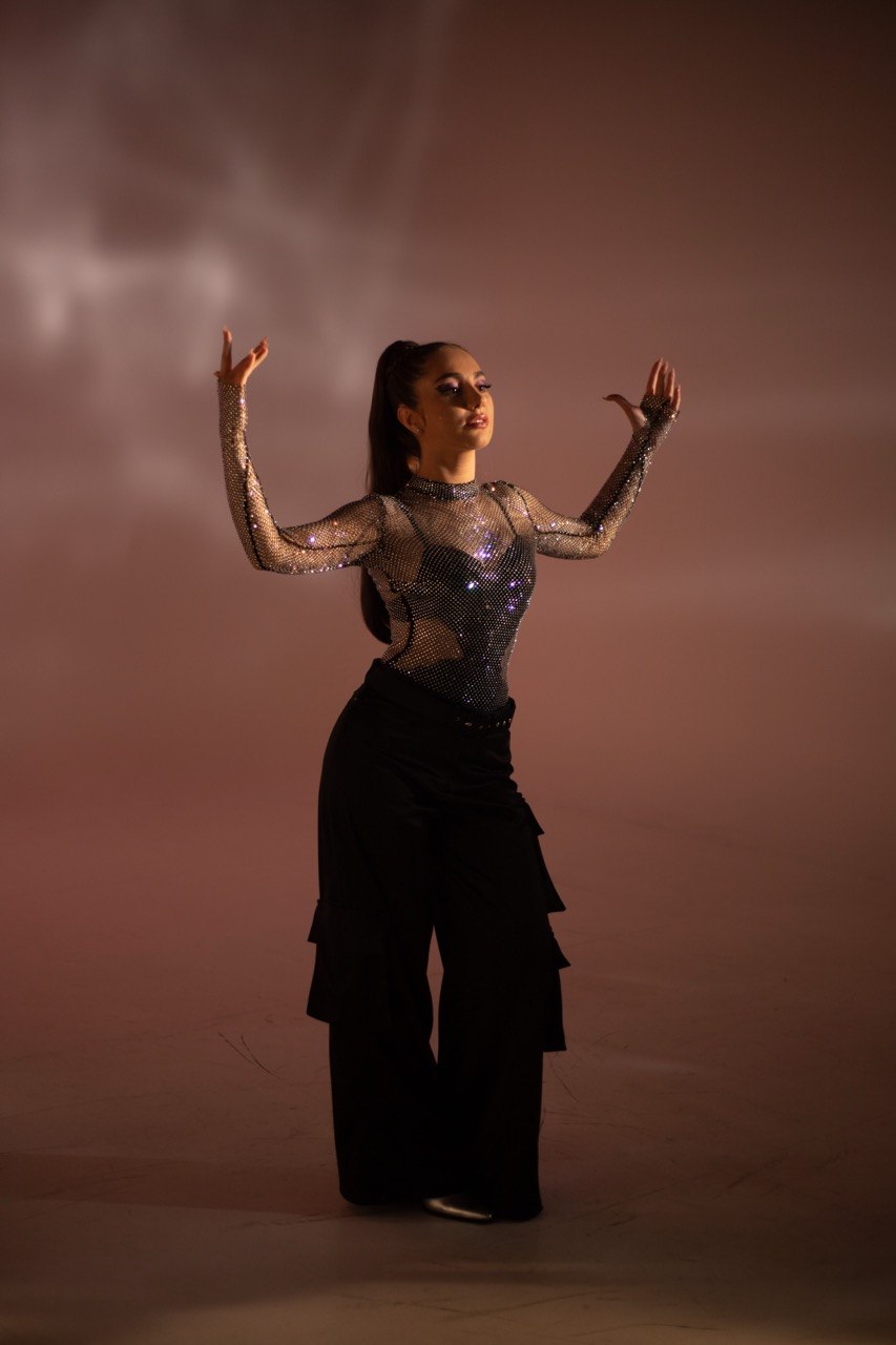 Person standing with arms raised, wearing a shiny top and wide pants, against a cloudy sky background.