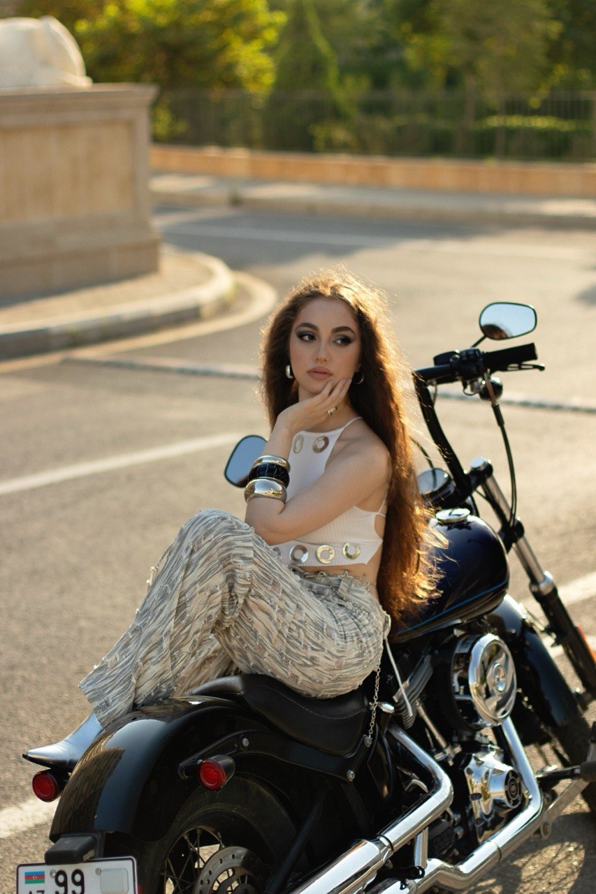 Young woman with long hair sitting on a black motorcycle on a street, with trees and a stone structure in the background.