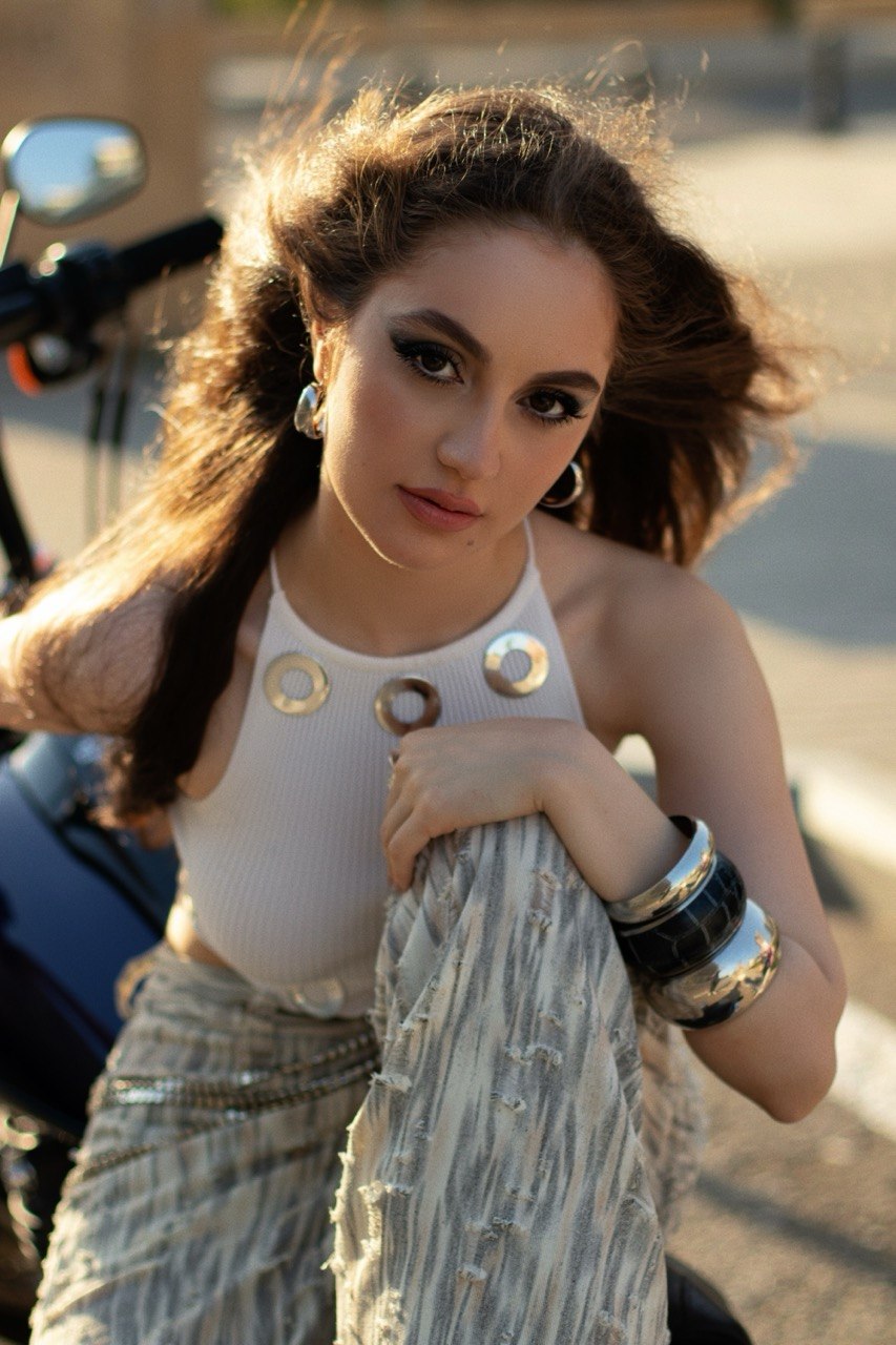 Young woman with long hair, wearing a white top with circular embellishments, sitting outdoors with sunlight, holding a fabric, wearing bracelets.