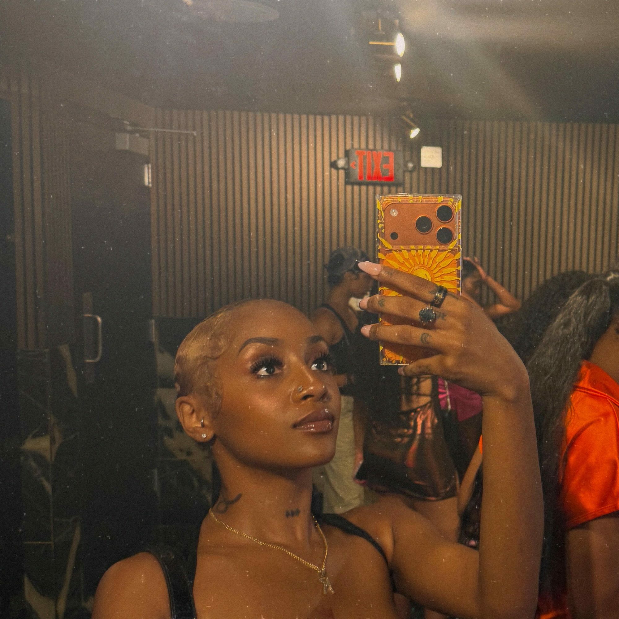 Woman with short hair taking a mirror selfie in a dimly lit room, holding a phone with a colorful case.
