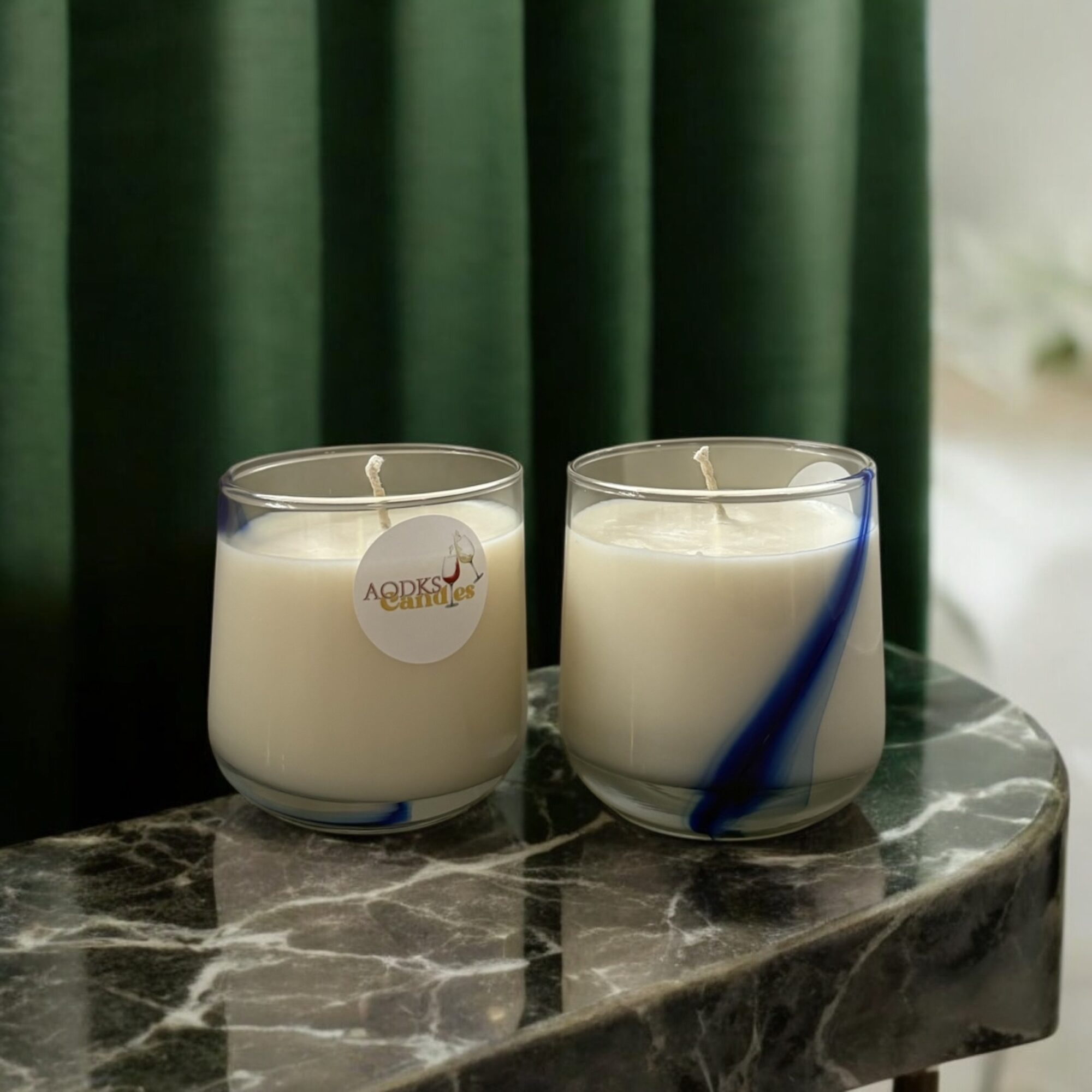 Two white candles in glass holders on a marble surface, with green curtains in the background.