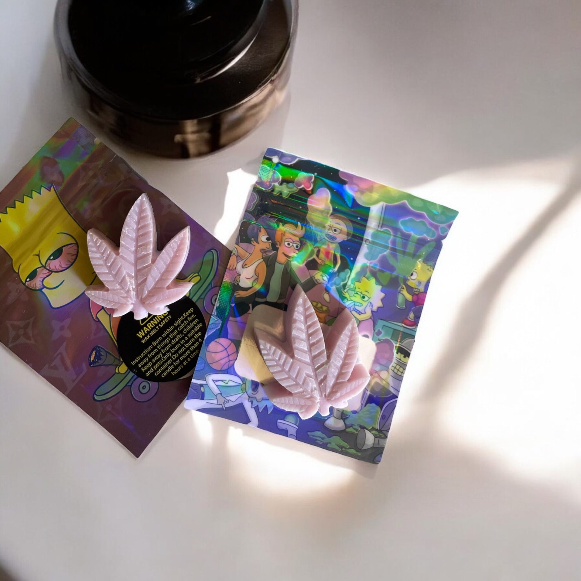 Two holographic cards with pink leaf-shaped objects and cartoon characters, placed on a white surface with a black container nearby.