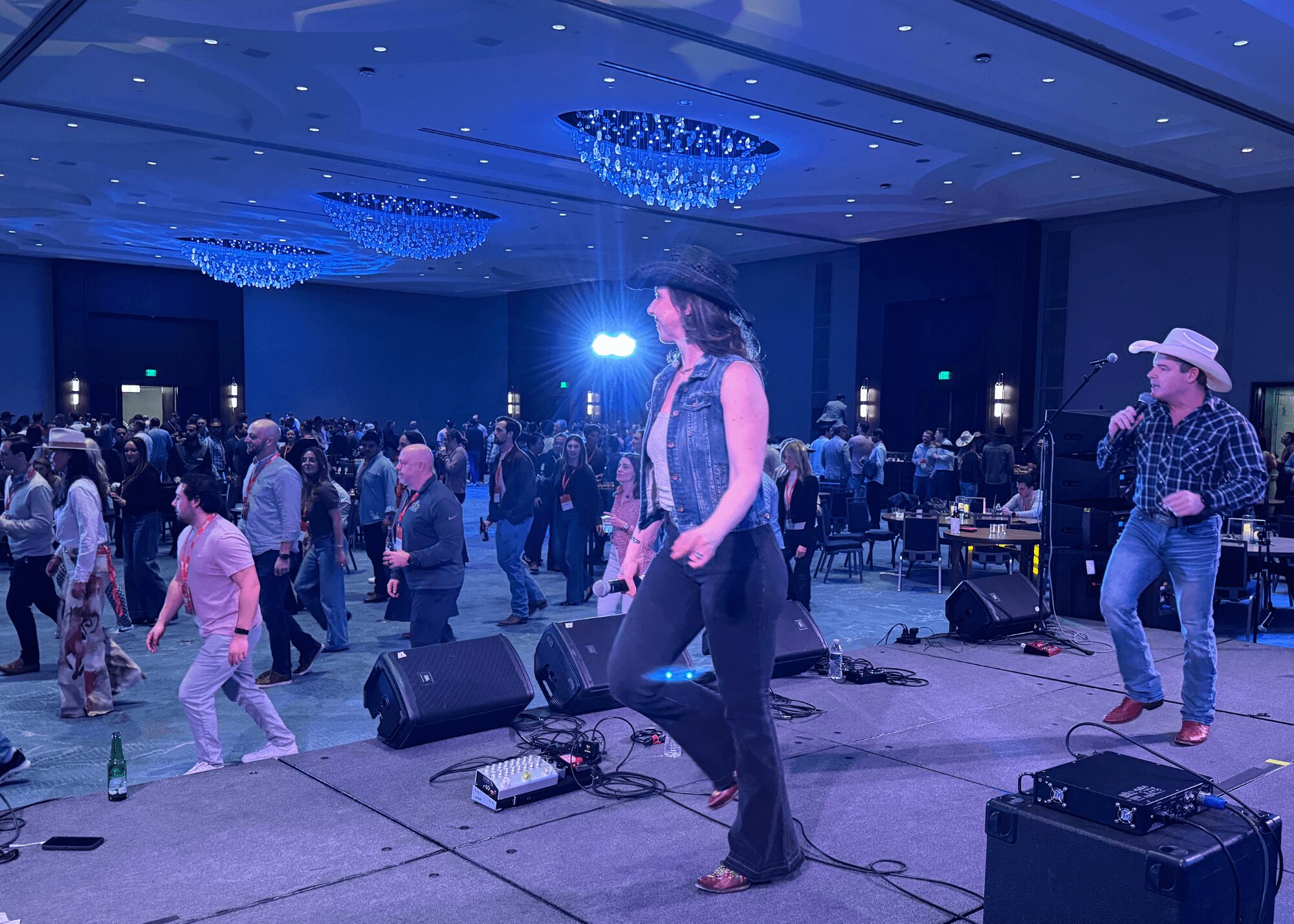 People dancing and a band performing on stage in a large indoor event space with blue lighting.