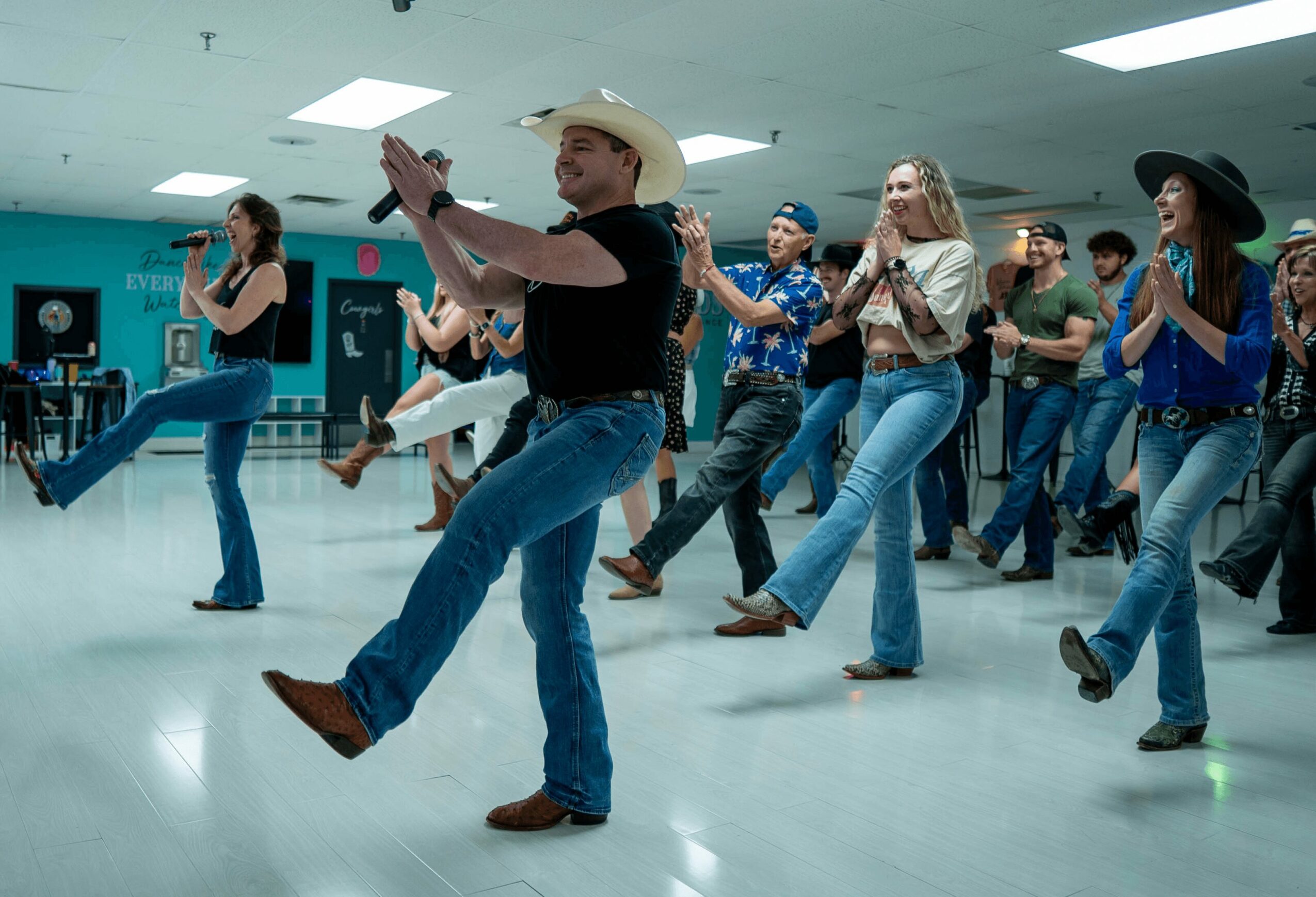 Group of people line dancing in a room, some wearing cowboy hats and jeans, with a man in the center kicking his leg up.