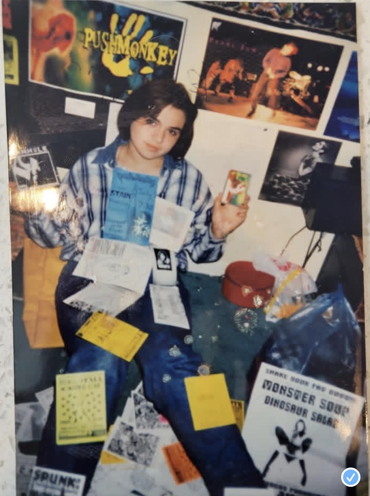 Person sitting at a table with various papers and items, posters on the wall behind, holding a phone, in an indoor setting.