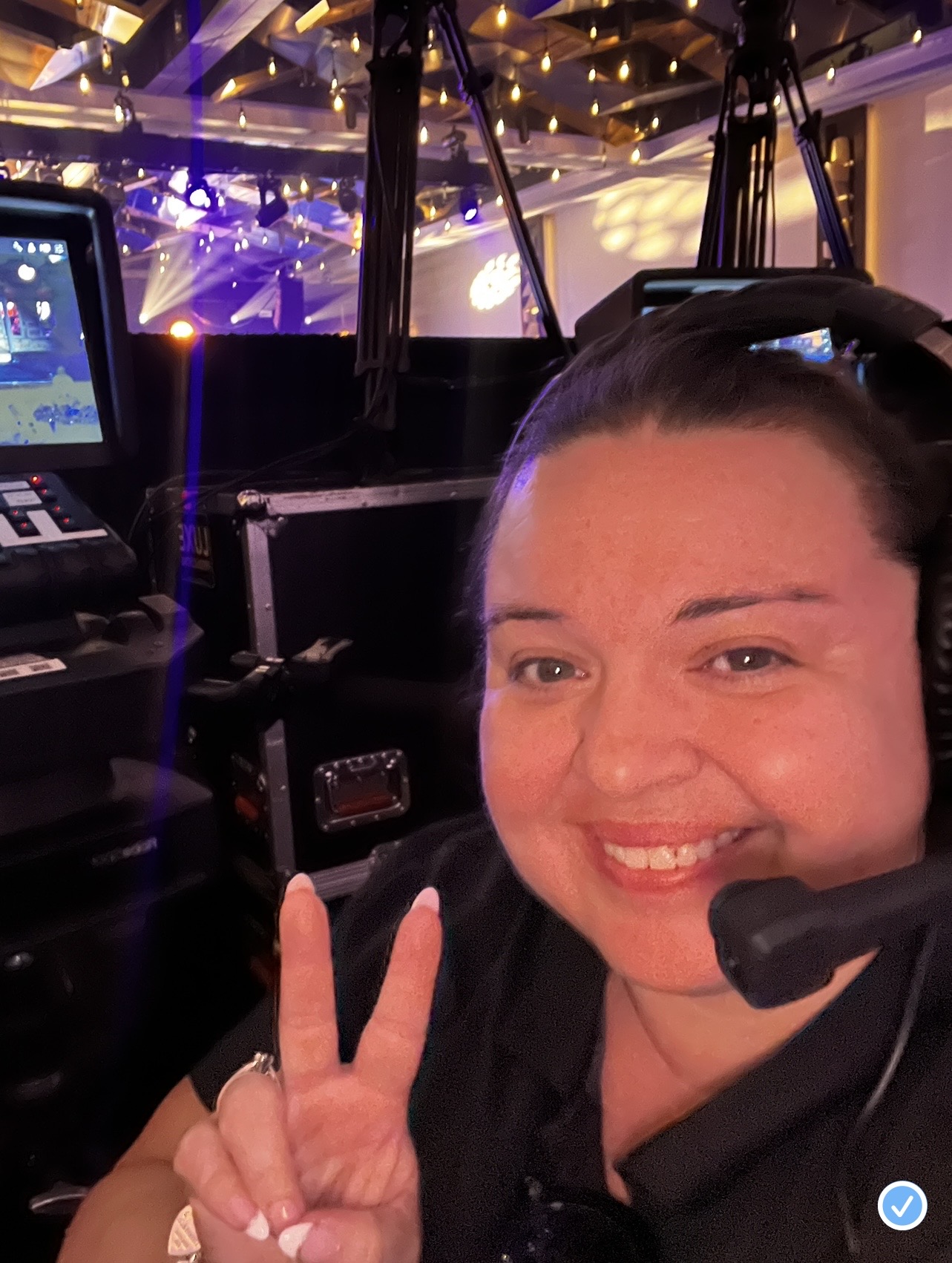 Smiling woman wearing a headset making a peace sign in a dimly lit event space with purple lighting.