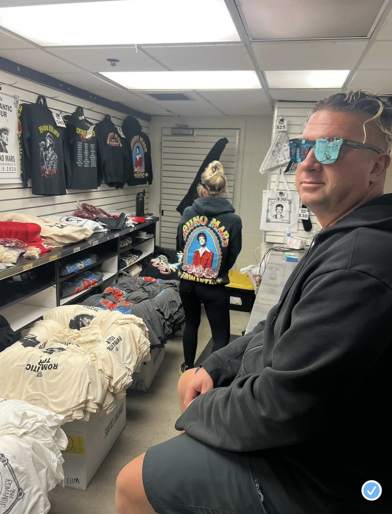 Man wearing sunglasses sitting in a store with clothing and merchandise on shelves and walls.