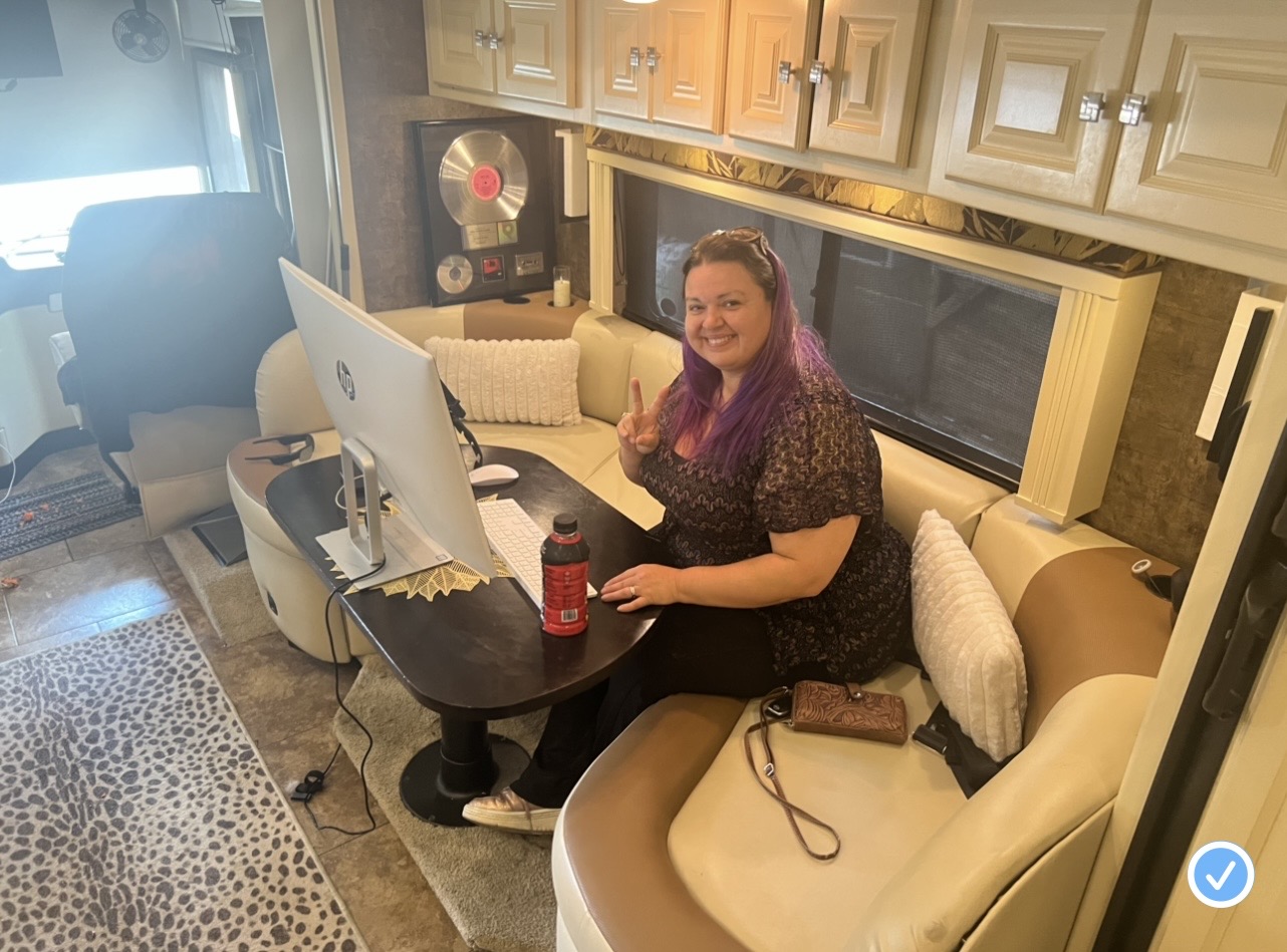 Woman with purple hair sitting at a table with a computer, smiling, in a cozy room with beige furniture and cabinets.