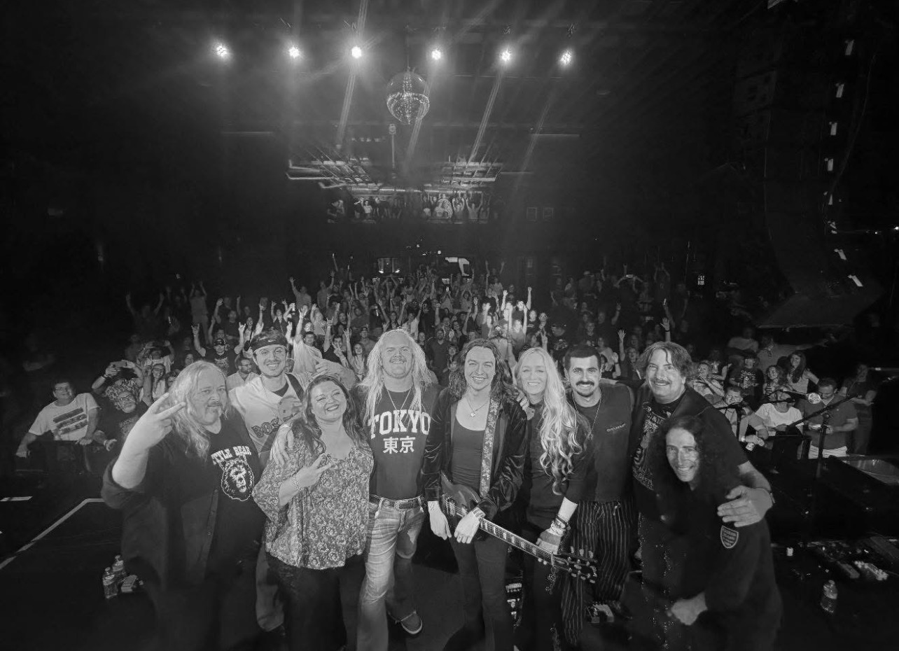 Group of people on stage in a concert venue with an audience and stage lights in the background.