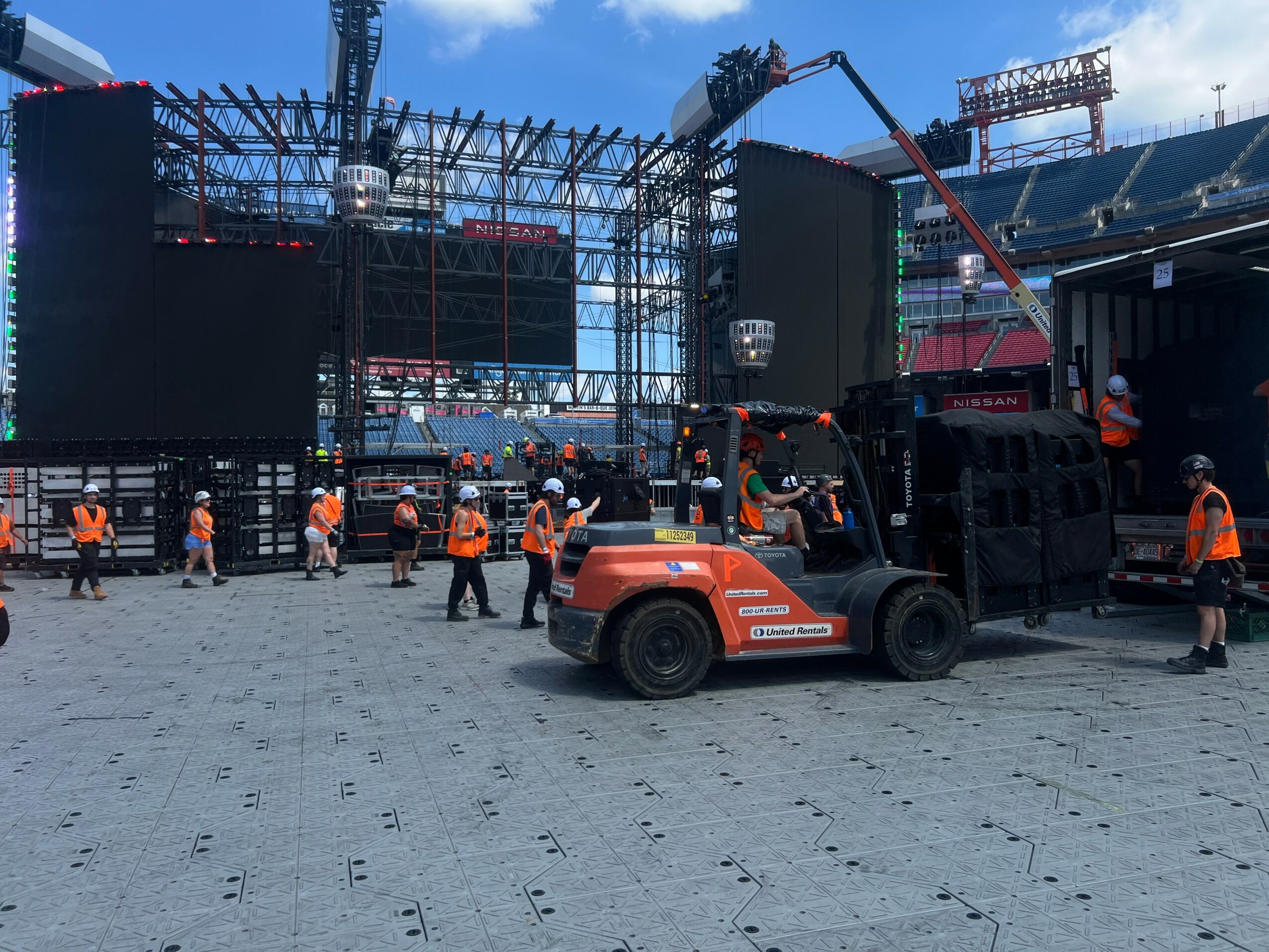 Construction workers set up stage equipment at a stadium with a large screen and scaffolding, blue sky overhead.