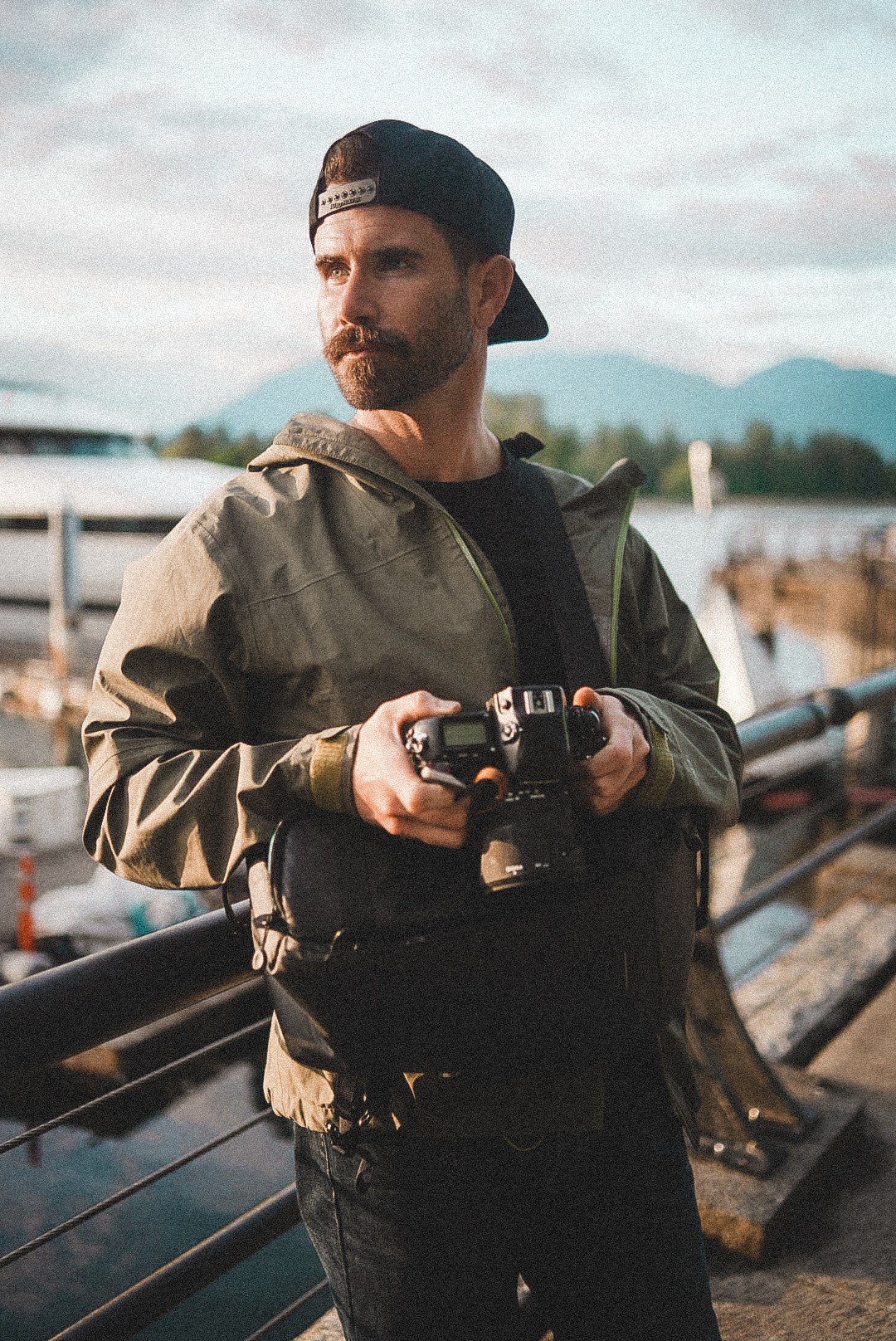 Man with beard wearing a black cap and jacket holding a camera, standing outdoors near water and boats.