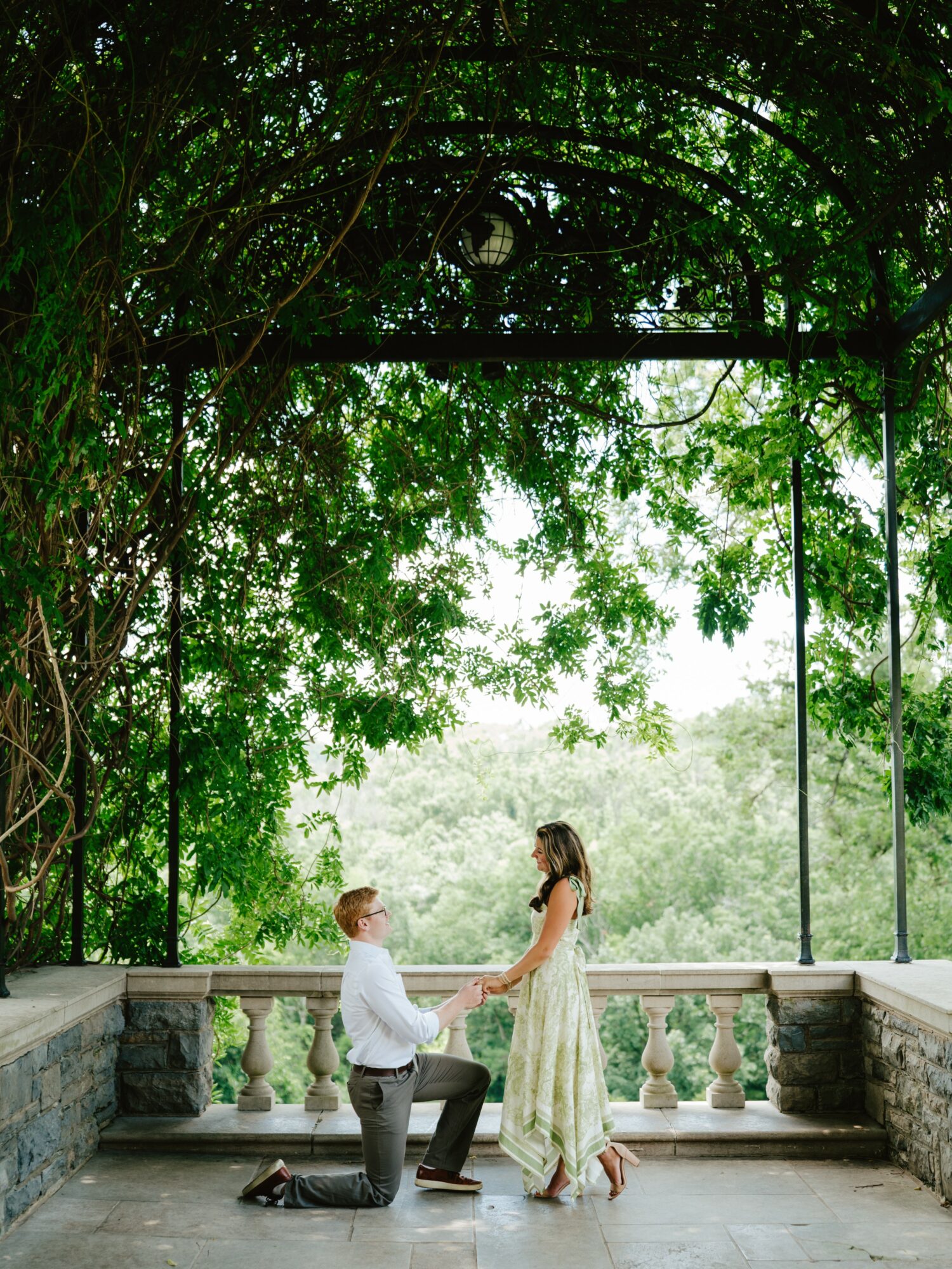 A man kneels holding a woman's hands on a balcony with trees overhead, bright sky in the background, and stone railing.