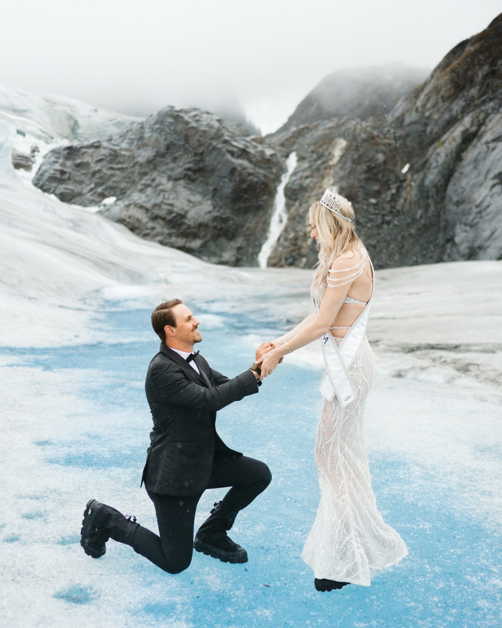 Man kneeling and holding hands with woman in a snowy landscape with mountains and waterfalls