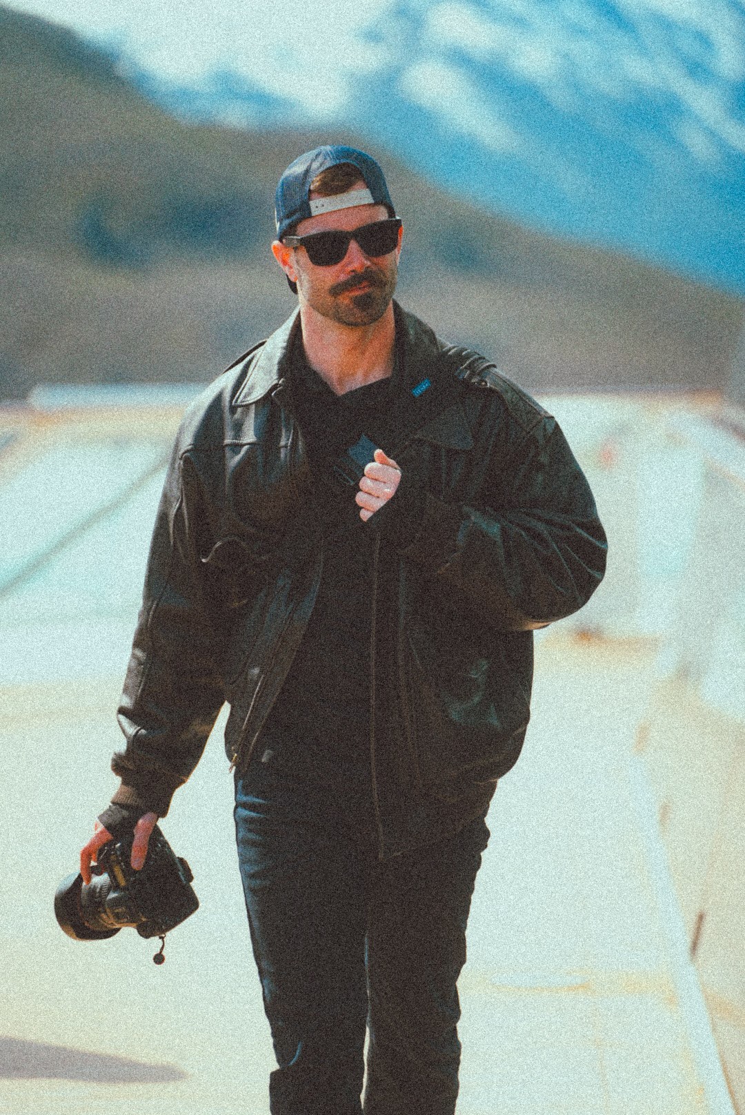 Man in black jacket and sunglasses walking outdoors with mountains in background.