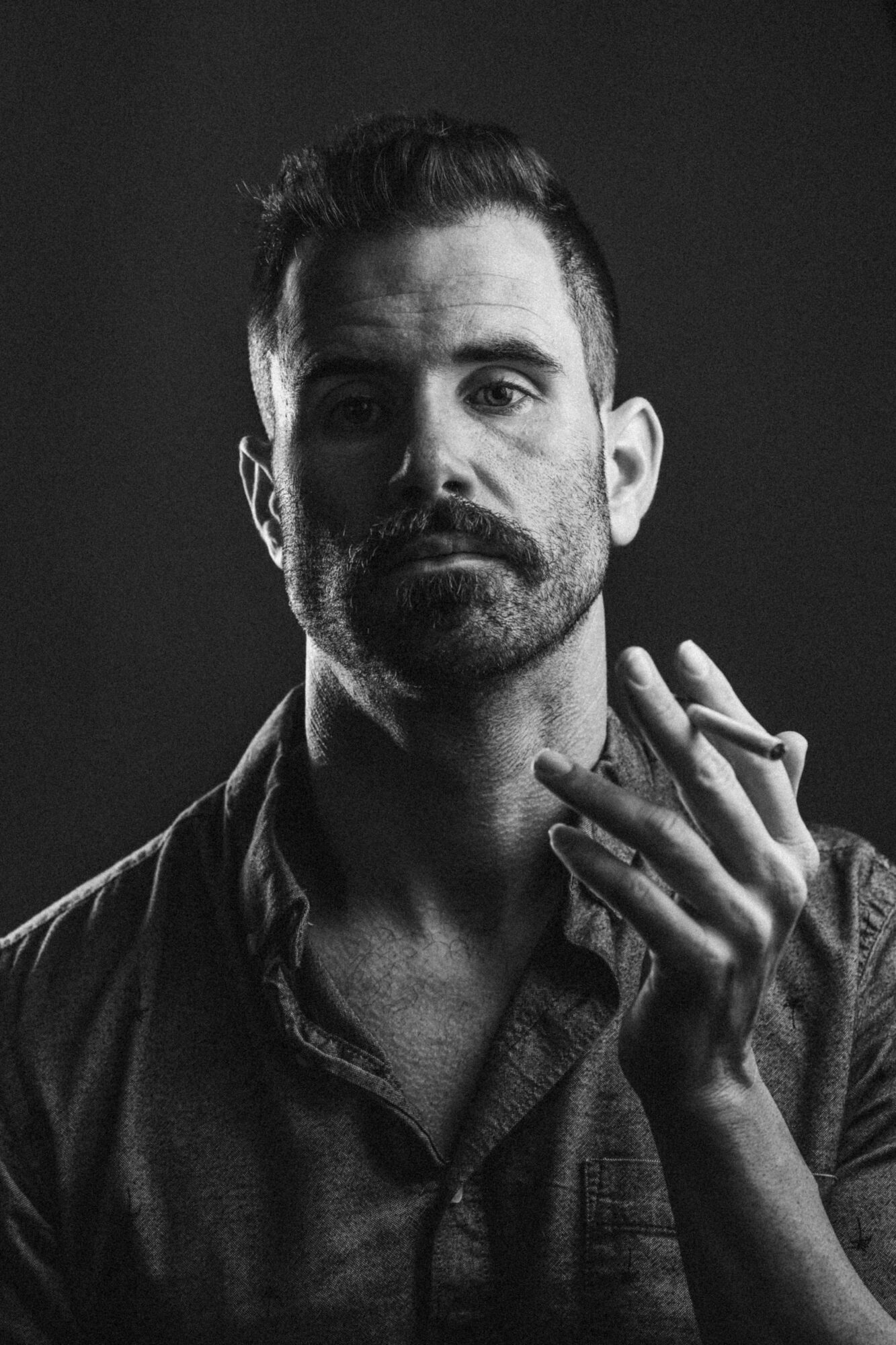 Black and white portrait of a man with a beard, short hair, wearing a collared shirt, looking at the camera, hand near face