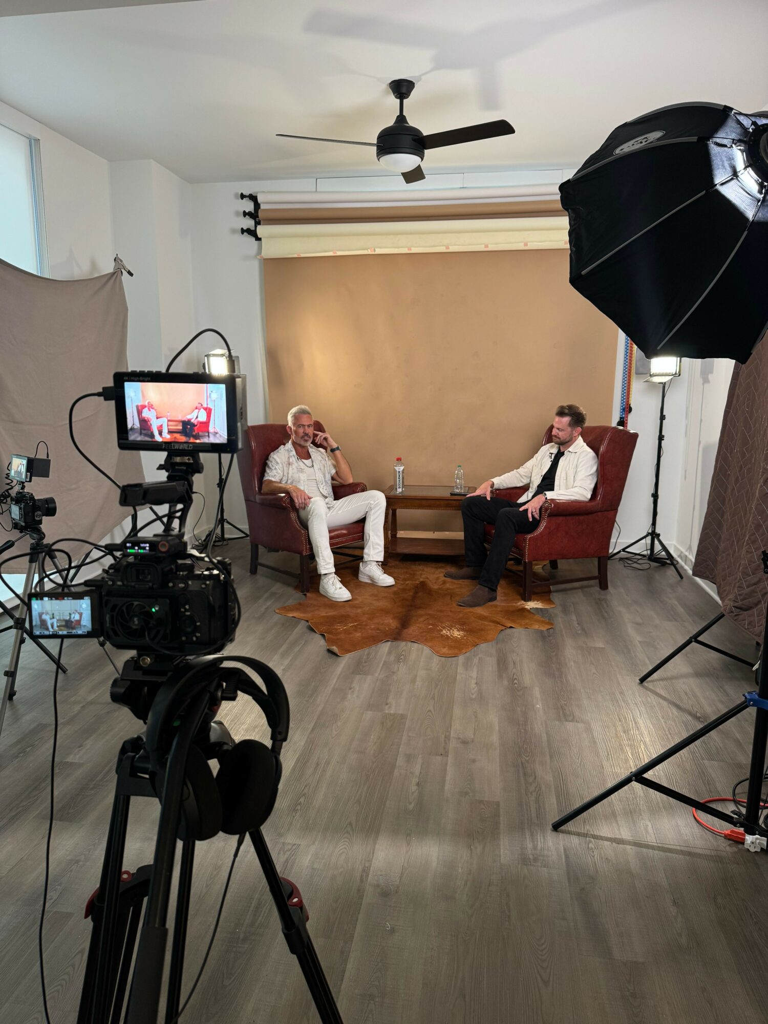 Two people sit on a couch in a photography studio with a backdrop, camera, and lighting equipment visible.