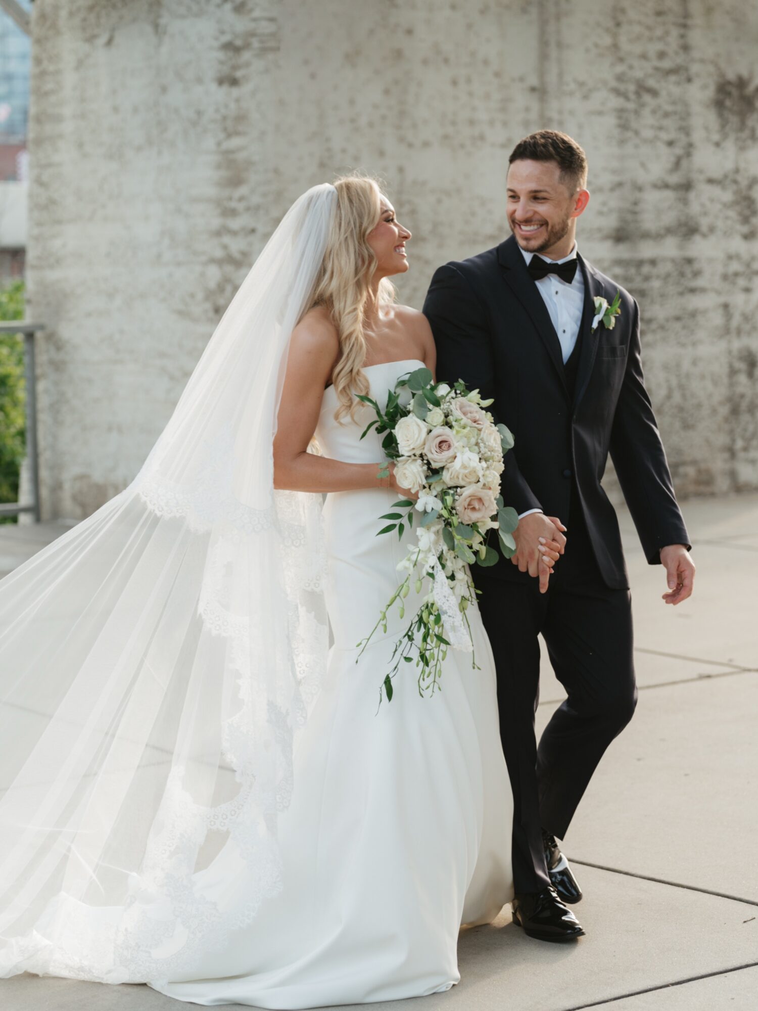Bride and groom holding hands, smiling, outdoors, bride in white gown with veil, groom in black tuxedo, wedding bouquet visible.