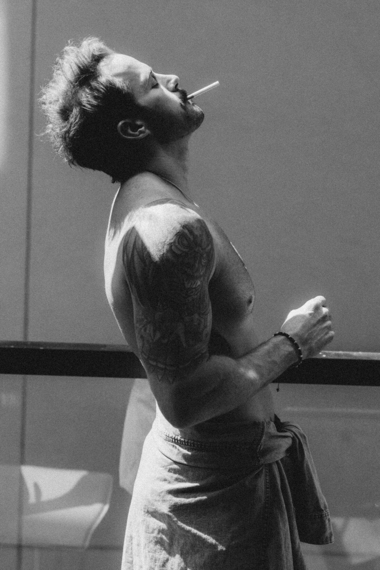 Side view of a shirtless person with tattoos smoking a cigarette, wearing pants, against a plain background.