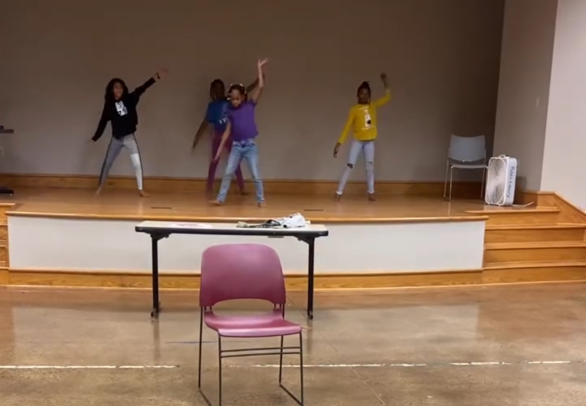 Four children dance on a stage with a wooden floor, a chair, and a table in front, in a room with plain walls.