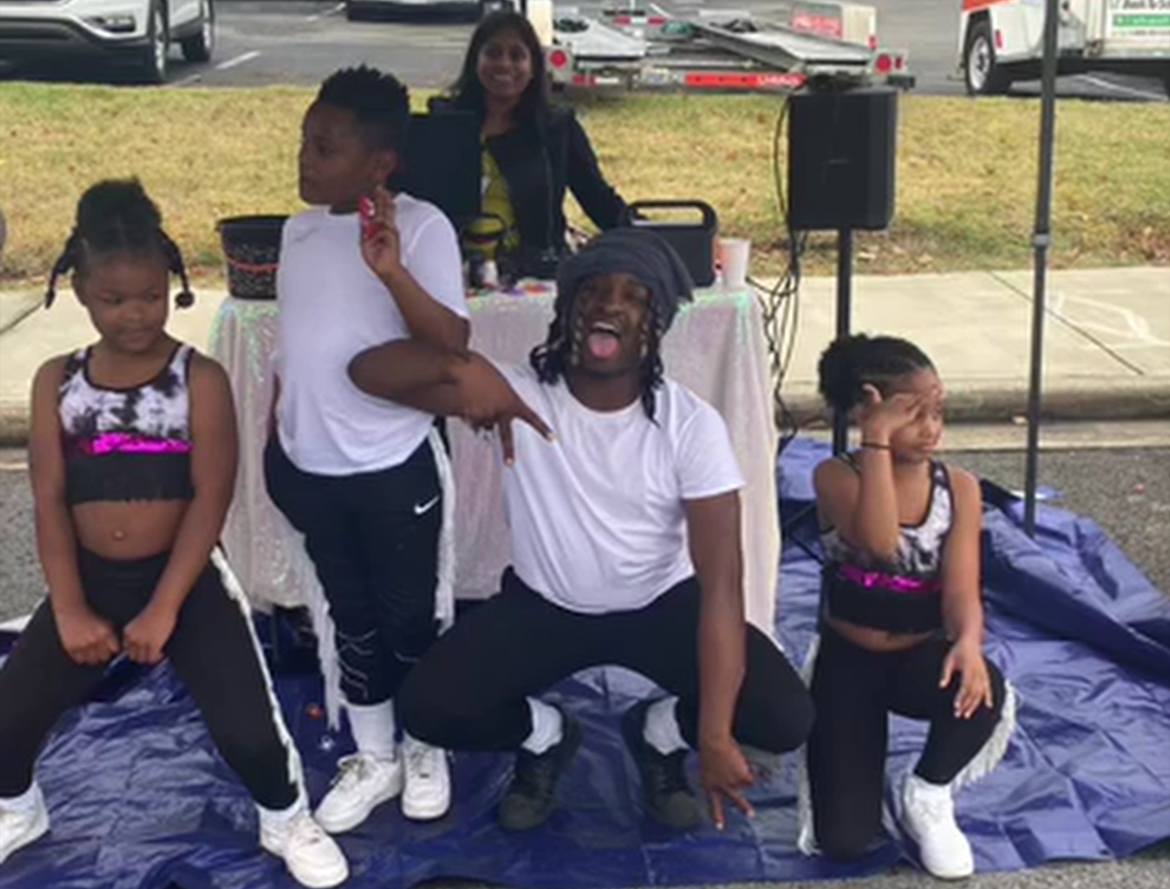 Group of children and an adult outdoors, with a woman sitting on the ground, children standing around, some covering their ears.