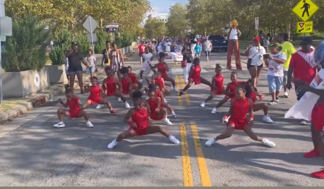 Group of children in red uniforms performing a synchronized dance or exercise on a street, with spectators and trees in the background.
