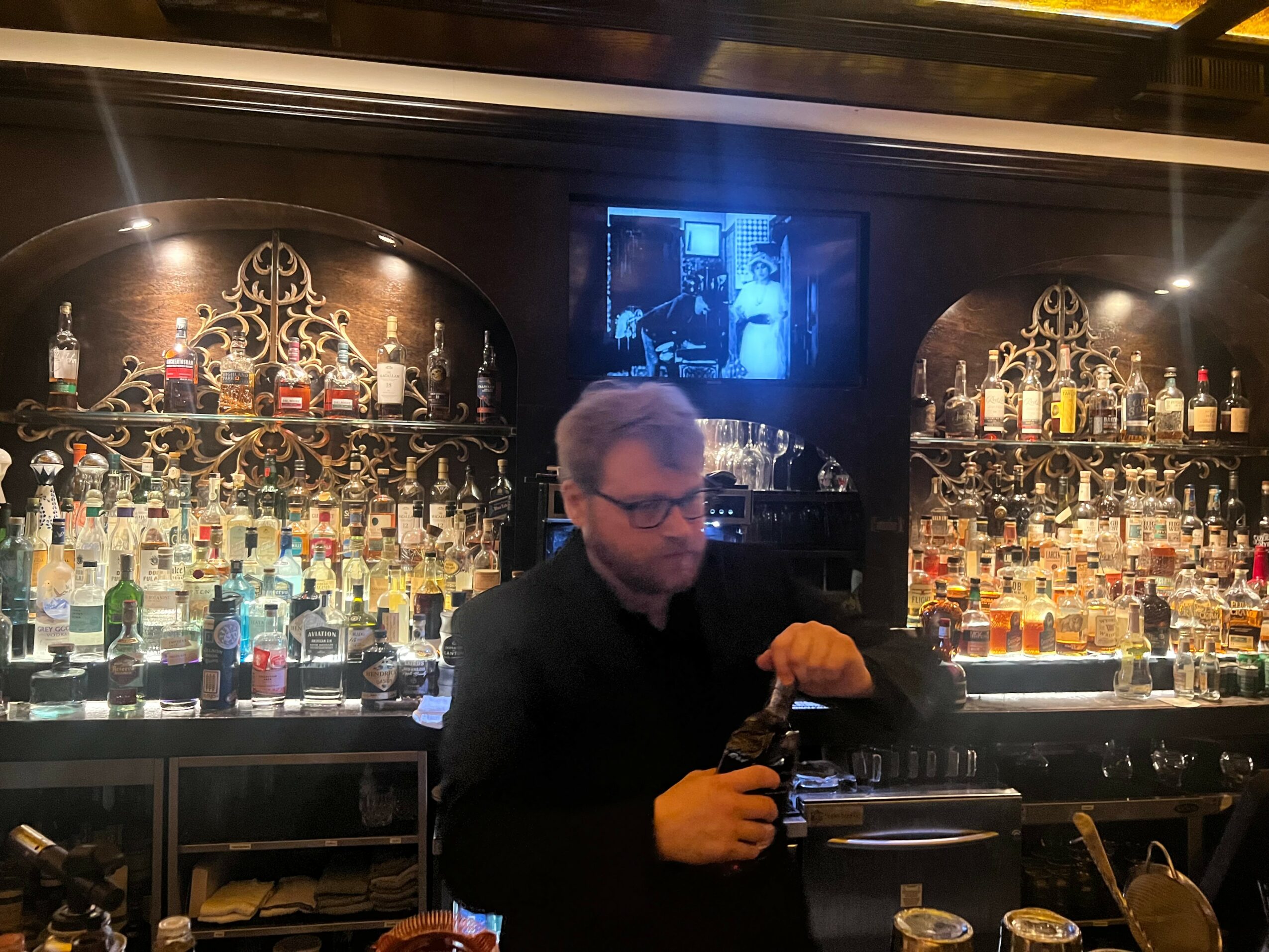 Man with glasses and beard behind a bar, surrounded by shelves of liquor bottles, in a dimly lit setting.