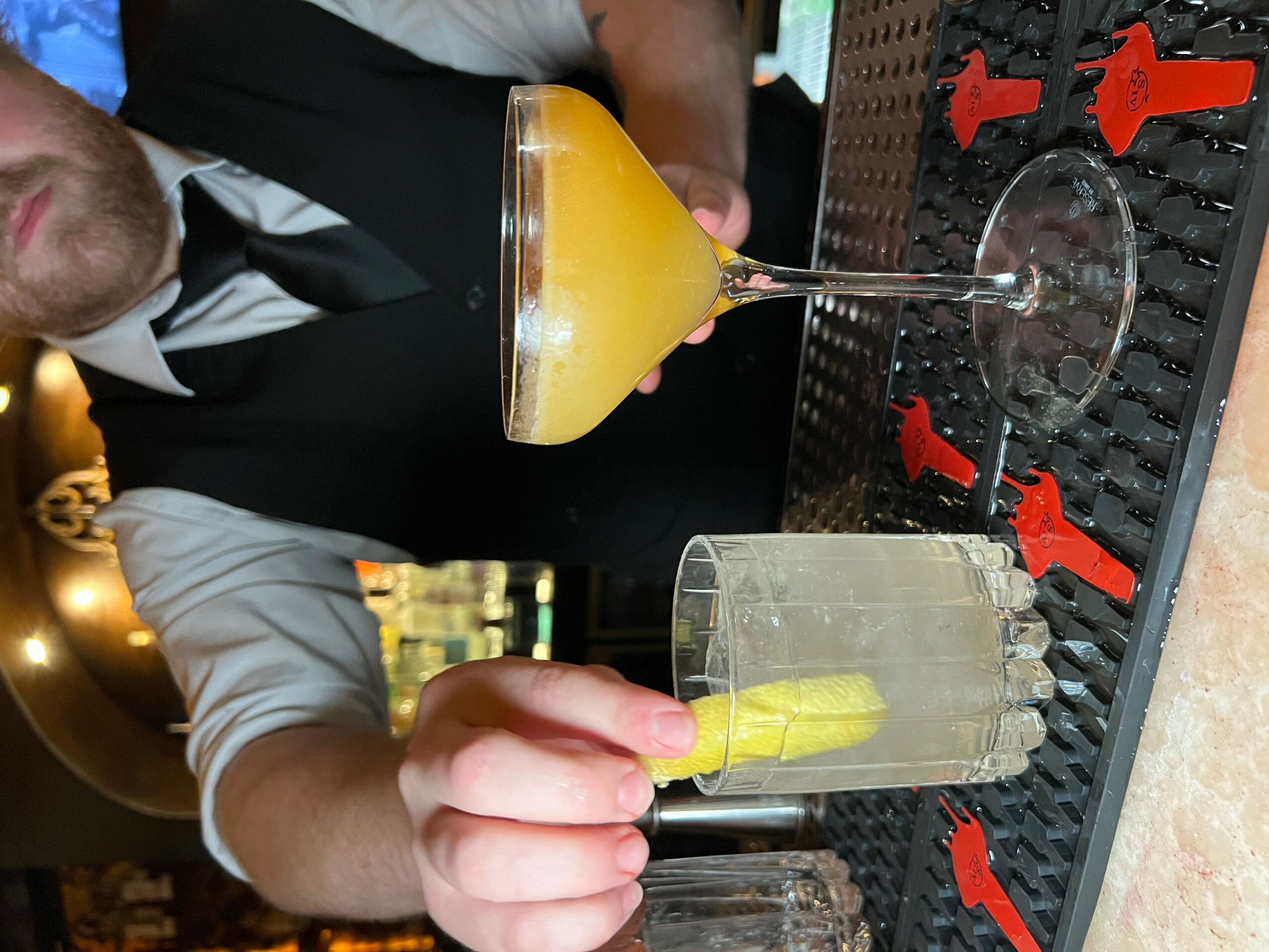 Person holding a yellow cocktail glass with a drink, sitting at a bar with a black rubber mat, red condiment bottles, and a glass of water.