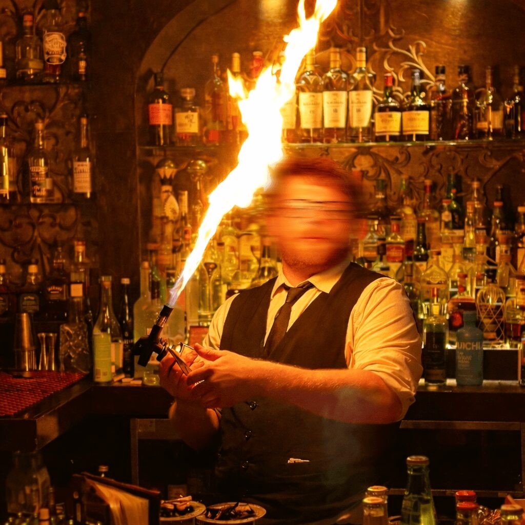 Bartender holding a flaming torch in a bar with shelves of liquor bottles behind him.