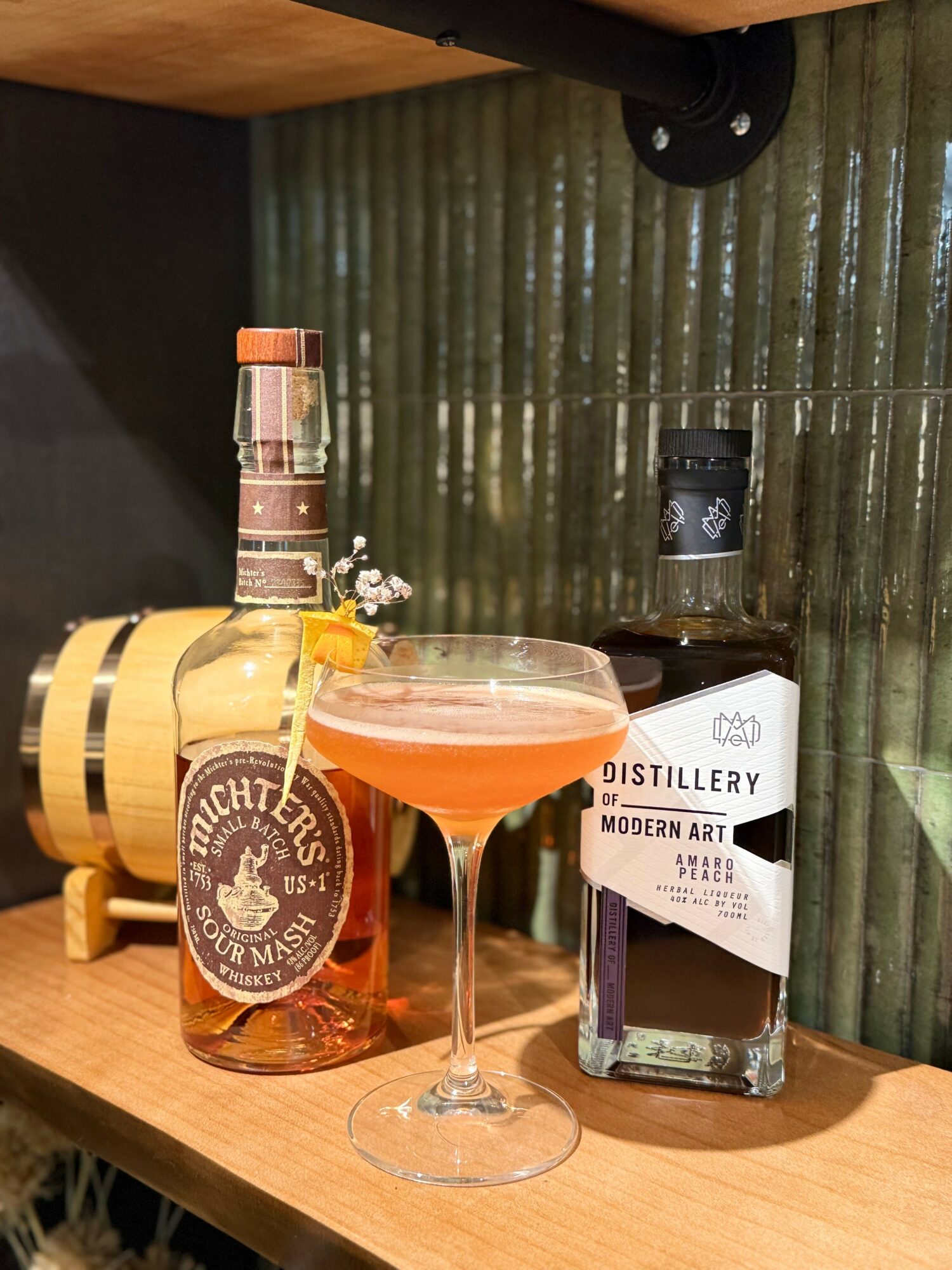 A cocktail glass with pink drink, a bottle labeled 'Distillery', and a small jar on a wooden shelf with green tiled background.