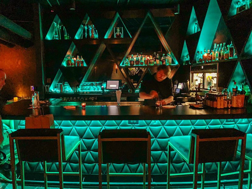 Bar with illuminated shelves and seating area, bartender preparing drinks, modern design with teal lighting.