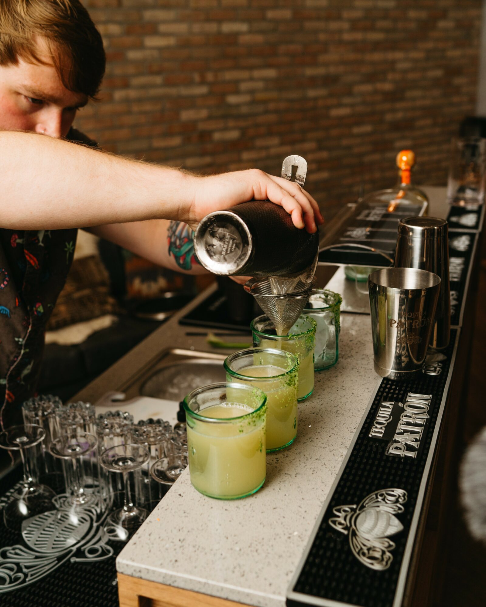 Person pours a dark liquid into glasses with green drinks on a bar counter, brick wall background.