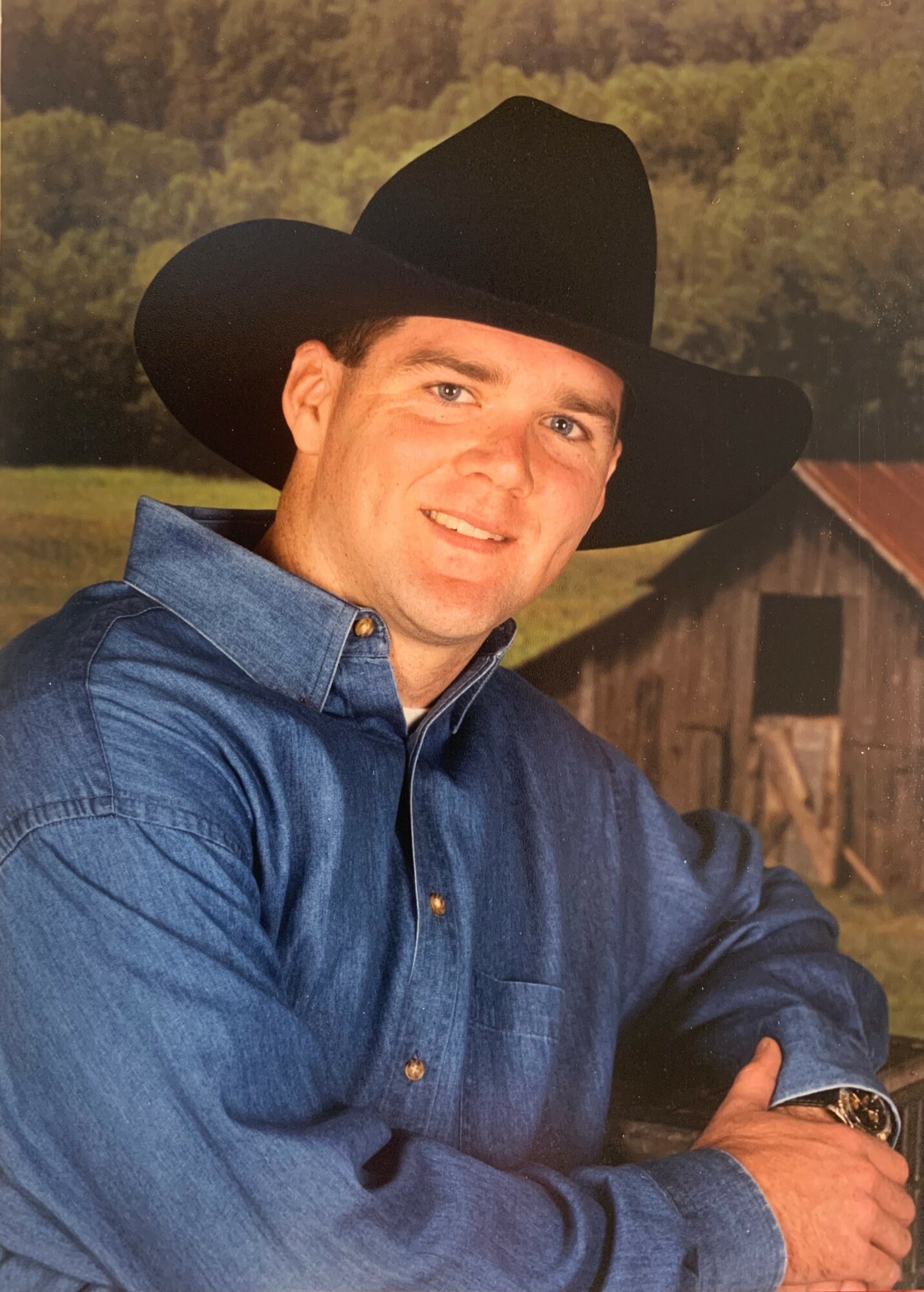 Man wearing a black cowboy hat and blue denim shirt, smiling outdoors with trees and a barn in background.
